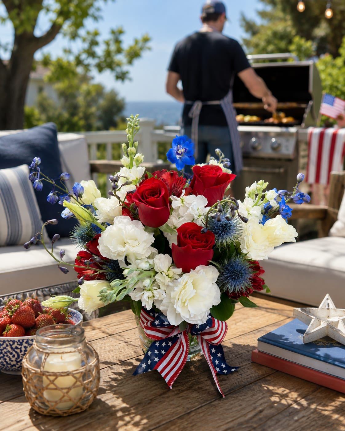 Patriotic Peninsula Blooms - Patriotic Peninsula Blooms is a fresh and festive celebration of summer featuring lush red roses, crisp white lisianthus, stock, and alstroemeria, accented with striking blue delphinium and thistle. Arranged in a 5" x 5" clear glass cylinder vase and adorned with a patriotic ribbon, this design offers a refined take on classic Fourth of July colors. Whether gracing a holiday table, welcoming guests to a summer gathering, or sent as a thoughtful gift, Patriotic Peninsula Blooms brings timeless seasonal charm with a distinctly coastal feel.