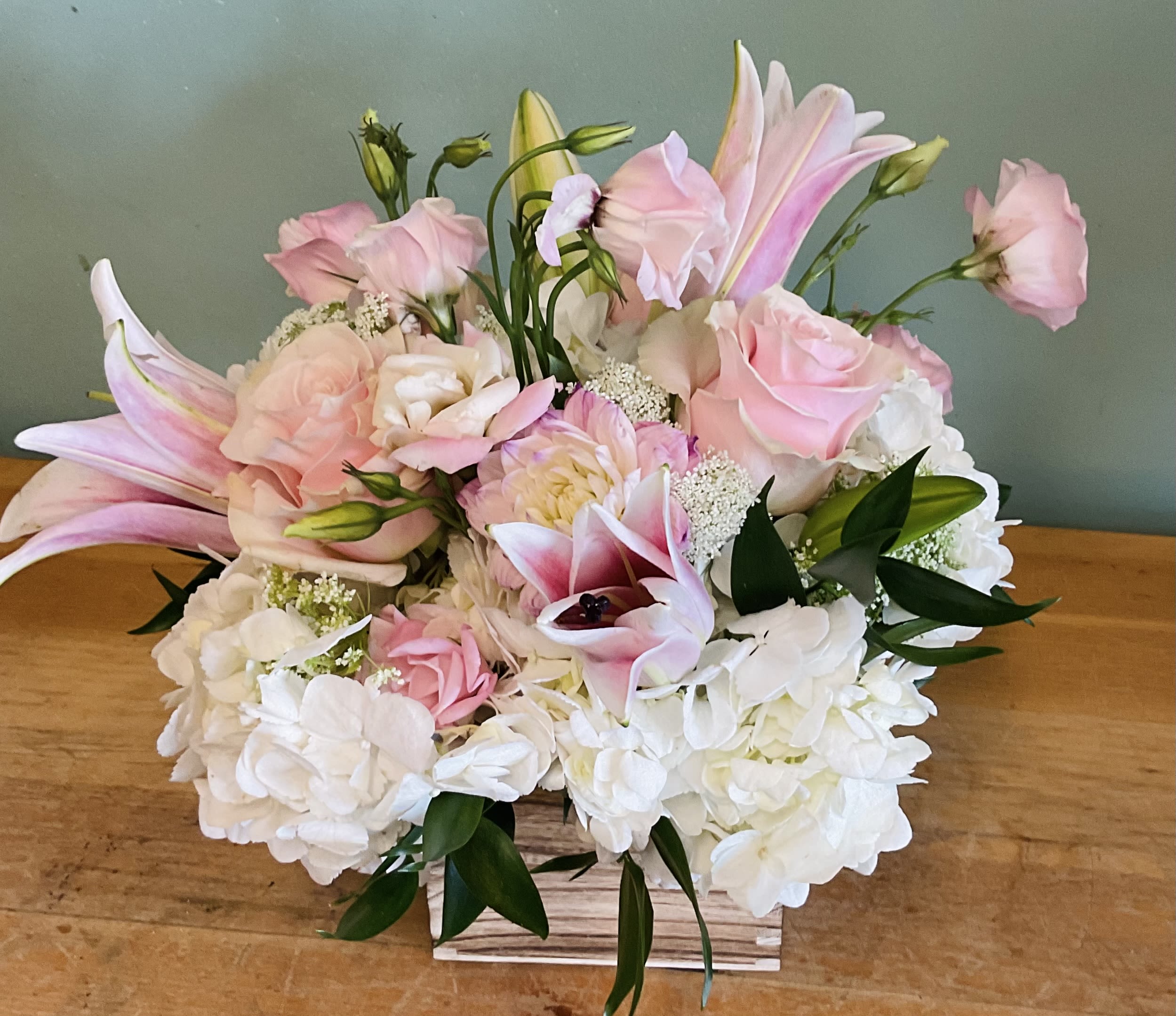  Blush Meadow Box - Soft pink lilies and roses mingle with creamy white hydrangeas and delicate lisianthus in a low rectangular wooden container-the perfect blush-toned flower arrangement for a birthday bouquet, spring celebration, or a thoughtful thank-you gift. The soft palette and rounded, garden-fresh silhouette create a gentle, romantic mood that will make the person you're thinking of feel cherished and seen.  This full, layered design pairs open blooms and buds for a textured, just-picked look. Pale pinks and whites are balanced by deep green foliage for contrast; the compact rectangular wooden box keeps the arrangement neat on a table or desk while adding a rustic, natural touch. Sized to fit small-to-medium surfaces, it arrives ready to display-just add fresh water and enjoy the fragrant lilies and long-lasting roses and hydrangeas.