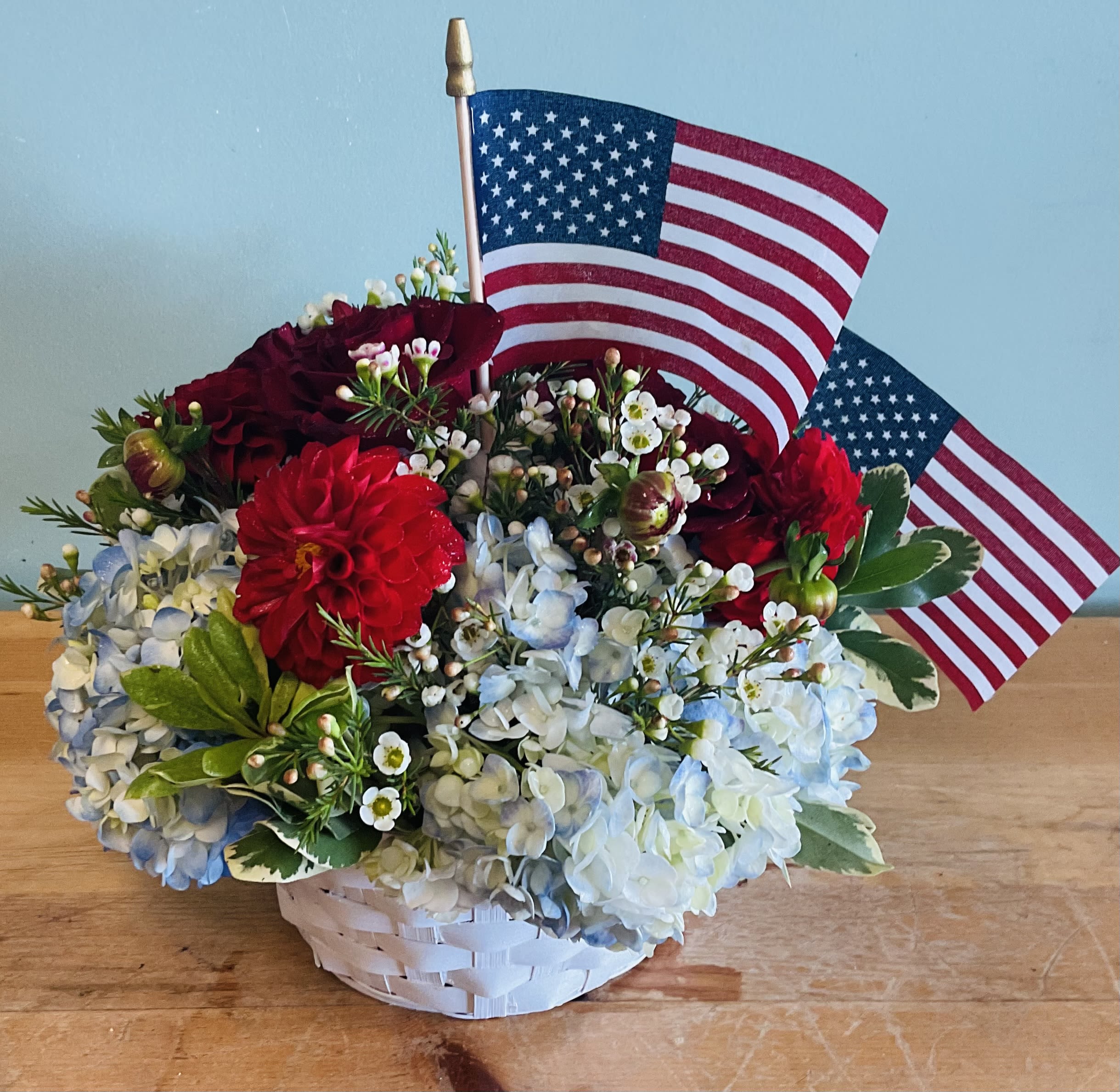 America 250 - A joyful sweep of red, white and blue-this patriotic flower arrangement is a bright way to celebrate a birthday, say "thinking of you," or mark Memorial Day and Fourth of July gatherings. The low, rounded basket design keeps the sentiment casual and homey, while two American flags and flag picks give the display a festive, proudly patriotic presence that makes the recipient feel noticed and celebrated.  Pale blue and white hydrangeas form a lush, cloud-like base, layered with vivid red dahlias and small white filler blooms for contrast and texture. Everything is arranged in a white wicker basket that sits compactly for tabletops or entry consoles; the dense, rounded composition reads full and abundant from every angle.  Perfect for sending "just because" appreciation to a neighbor, veteran, or family member, this arrangement brings seasonal spirit without feeling limited to holiday use. Order with standard delivery timing-refresh vase water daily and keep blooms out of direct afternoon sun to help the flowers look their best for several days.