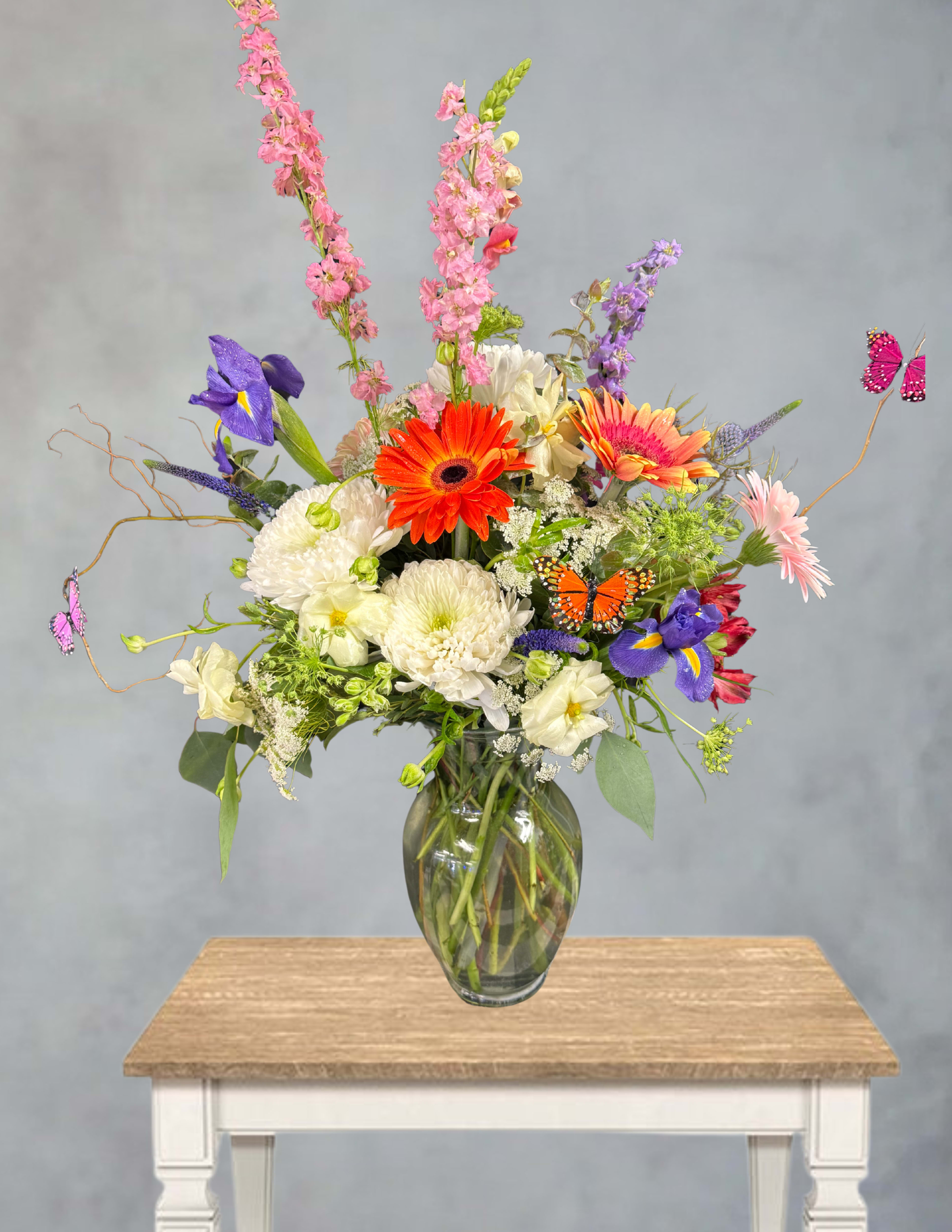 Wild as a Butterfly  - Bright orange gerbera daisies leap forward in this Wild as a Butterfly flower arrangement, paired with purple iris, soft pink snapdragon spikes, and crisp white chrysanthemums to create a playful birthday bouquet or springtime gift. The airy, asymmetrical design and whimsical butterfly picks make it an ideal choice to celebrate a cheerful moment-she'll feel noticed and uplifted the minute she sees those vibrant colors and movement.  Arranged in a clear glass vase, the mix of gerbera daisies, iris, chrysanthemums and snapdragons extends outward and upward for a garden-fresh silhouette that reads lively from every angle. The palette of orange, purple, pink and white offers strong visual contrast while keeping a light, celebratory mood; decorative butterfly picks add a charming finishing touch. Perfect for birthdays, a "thinking of you" surprise, or any spring occasion, this arrangement arrives ready to display-no extra fussy care required beyond fresh water and keeping blooms out of direct heat for longer enjoyment.