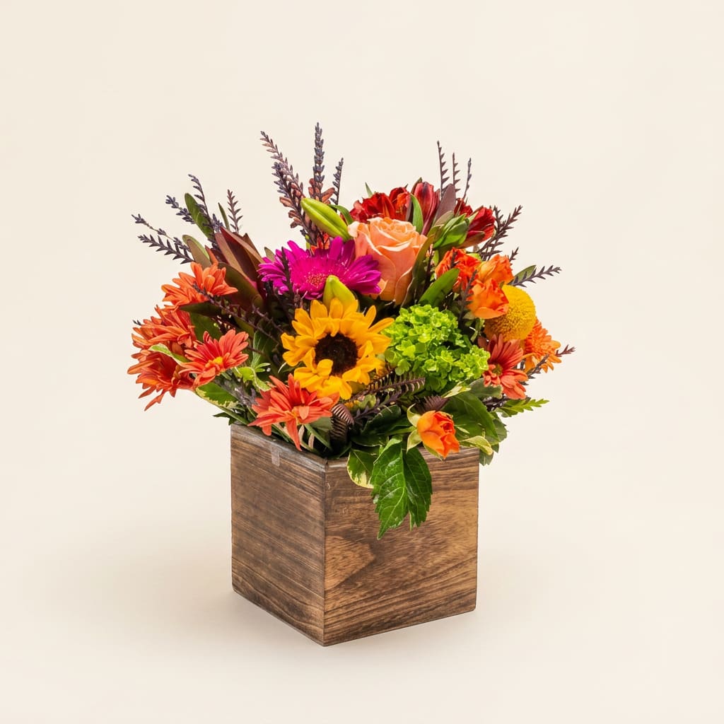 Warm Harvest - A sunflower-like focal bloom and a peach rose rising behind it create an instantly cheerful centerpiece-perfect as a fall birthday bouquet or a warm-season gift. This flower arrangement for seasonal gifting and everyday celebrations uses mixed-color roses, sunflowers, gerbera daisies, chrysanthemums, and complementary blooms to deliver a vibrant, autumn-leaning palette that reads festive without being formal. The overall effect is lively and welcoming, the kind of bouquet that will make the person you're thinking of feel genuinely seen.  Tightly layered orange, pink, yellow, and red flowers fill a square wooden cube vase for a full, energetic look; upright stems and varied textures-soft rose petals, bold daisy faces, and fluffy mums-add visual depth from every angle. The rustic wood container brings a handcrafted warmth that suits a kitchen table, office desk, or entryway. Sized to sit as a noticeable yet not overwhelming display, this arrangement arrives ready to enjoy.  Ideal for saying thanks, celebrating a birthday, or brightening someone's day, Warm Harvest feels seasonal and versatile-appropriate for hosts, colleagues, or a close friend. To keep blooms looking fresh, place in a cool spot away from direct sun and refresh the water every few days; change the water and trim stems slightly if you'd like to extend vase life.