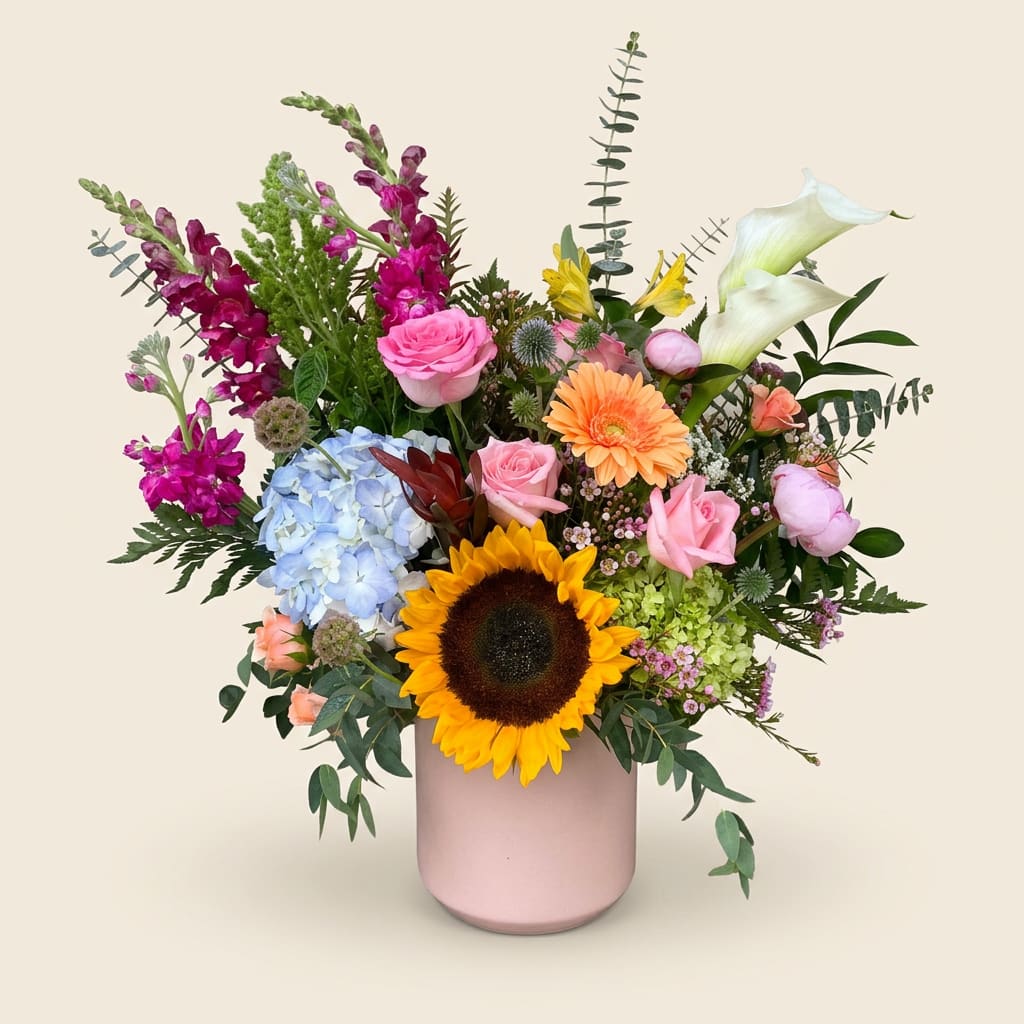 Wild Contrast - A single, bold sunflower anchors a lively mixed-color bouquet that feels like a sunlit garden in bloom-perfect as a birthday bouquet, a cheerful thank-you arrangement, or an everyday gift to brighten someone's day. The arrangement's asymmetrical, airy silhouette and upbeat palette of yellow, pink, purple, blue, and white make the recipient feel genuinely celebrated and noticed, whether you're sending birthday wishes or a spontaneous pick-me-up.  This flower arrangement includes a large sunflower, pale blue hydrangea, pink roses, white calla lilies, vivid magenta snapdragons, alstroemeria, daisies, and complementary foliage, artfully gathered in a rounded but fresh shape. Stems extend upward and outward for a natural, garden-style look; the flowers arrive nestled in a pink ceramic vase that adds a soft, modern touch and makes the gift display-ready.  Ideal for brightening a kitchen table, office desk, or bedside, this piece is designed to feel abundant without being overly formal-she'll feel cheered and seen the moment it arrives. Vase care is simple: trim stems and refresh water every few days to keep blooms lively; available in the pictured arrangement size.