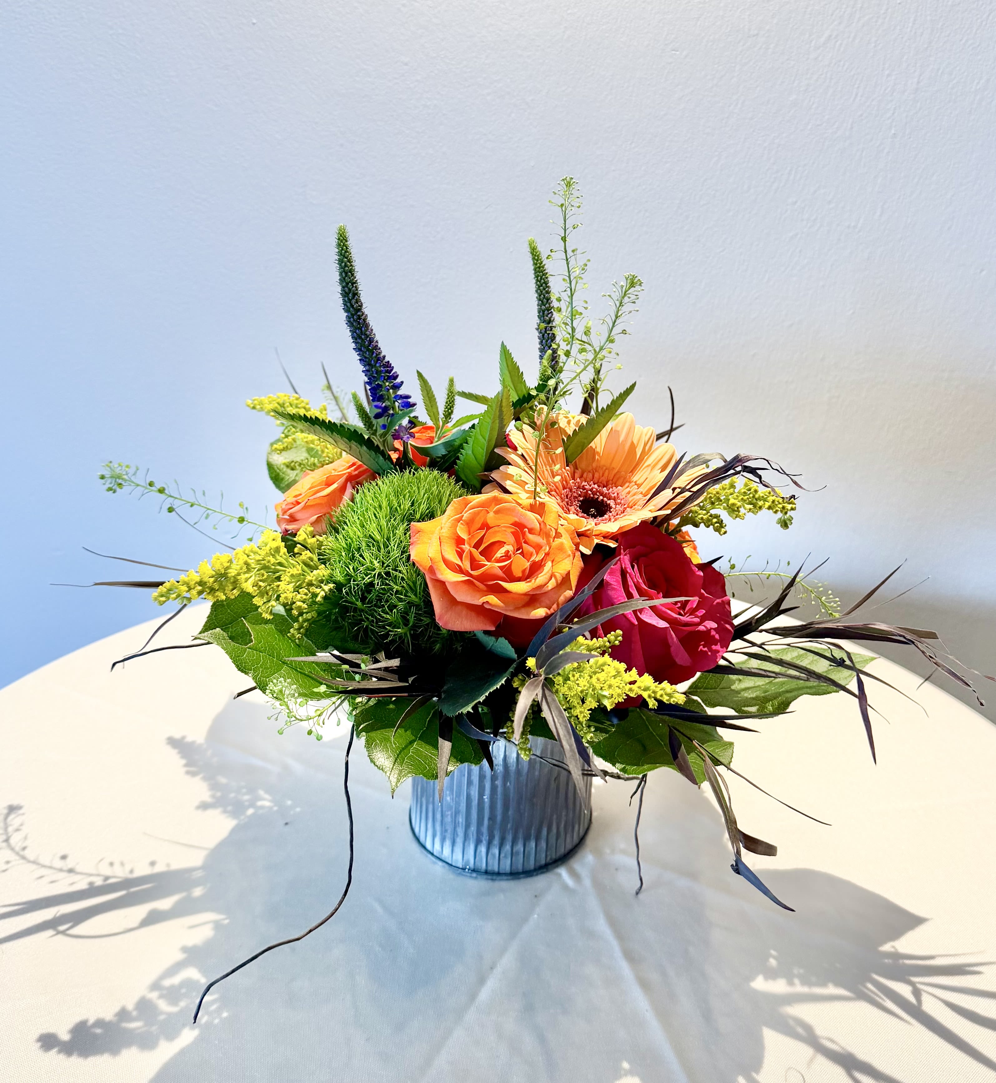 Summer Song - A joyful mix of vibrant blooms and lush textures, Summer Song captures the beauty of a summer garden in full bloom. Featuring premium roses, cheerful gerbera daisy, vibrant green trick dianthus, graceful veronica, airy accents, and seasonal greenery, this arrangement is designed in a short modern metal container for a fresh, contemporary look. Perfect for birthdays, celebrations, thank-you gifts, or simply brightening someone's day.