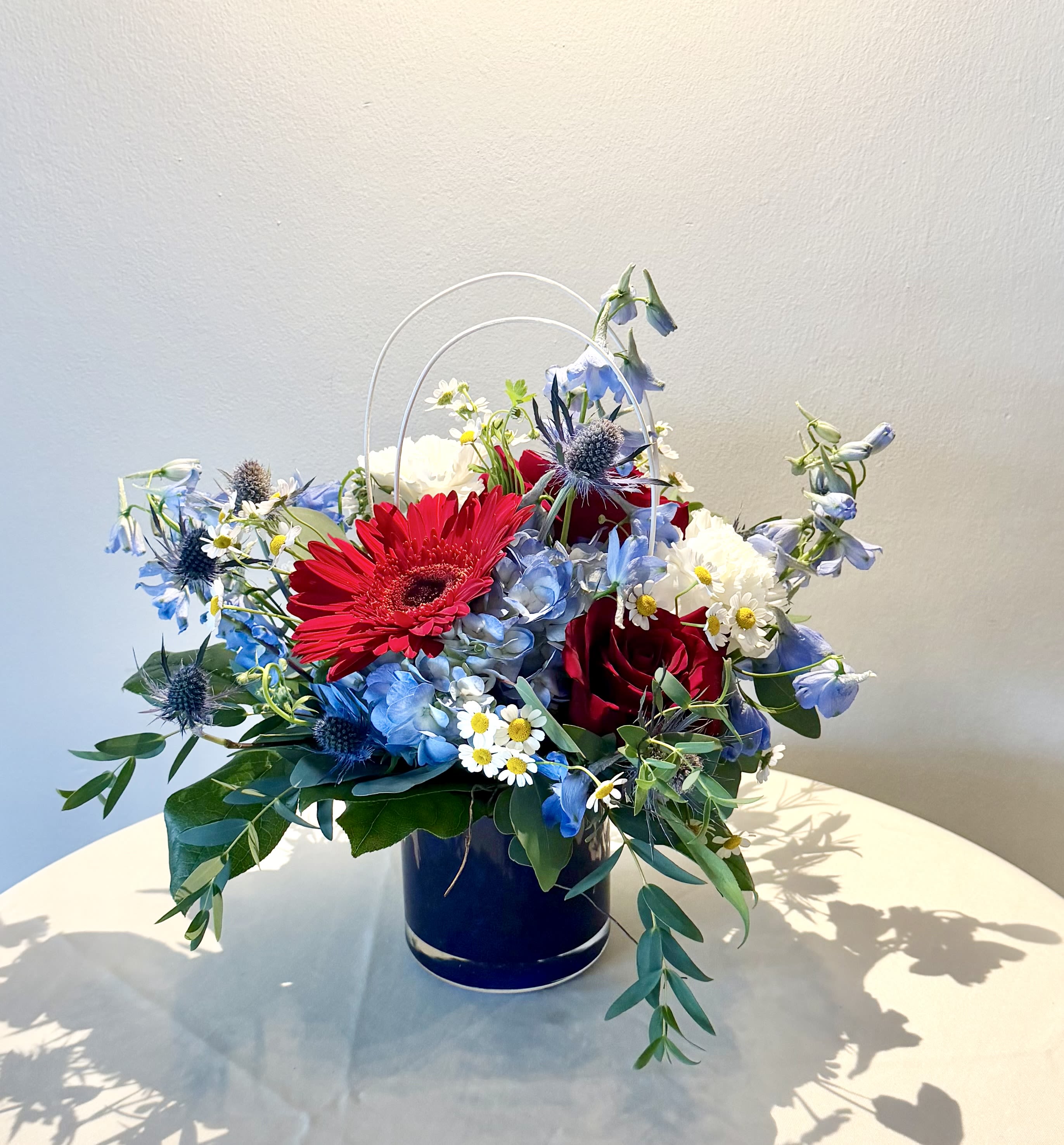 Fireworks - Celebrate the spirit of summer with Fireworks, an elegant arrangement inspired by the colors of the Fourth of July. Rich red roses and vibrant gerbera daisies are nestled among soft blue hydrangea, graceful delphinium, delicate chamomile daisies, white carnations, and textured thistle, all accented with lush seasonal greenery. Designed in a modern blue keepsake container, this festive arrangement is perfect for holiday gatherings, hostess gifts, barbecues, or simply adding a touch of patriotic charm to any home.