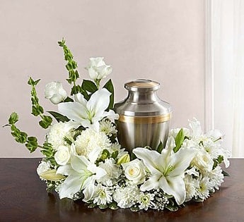 White lilies, roses, and mums arranged in a ring around a silver urn on a dark table.