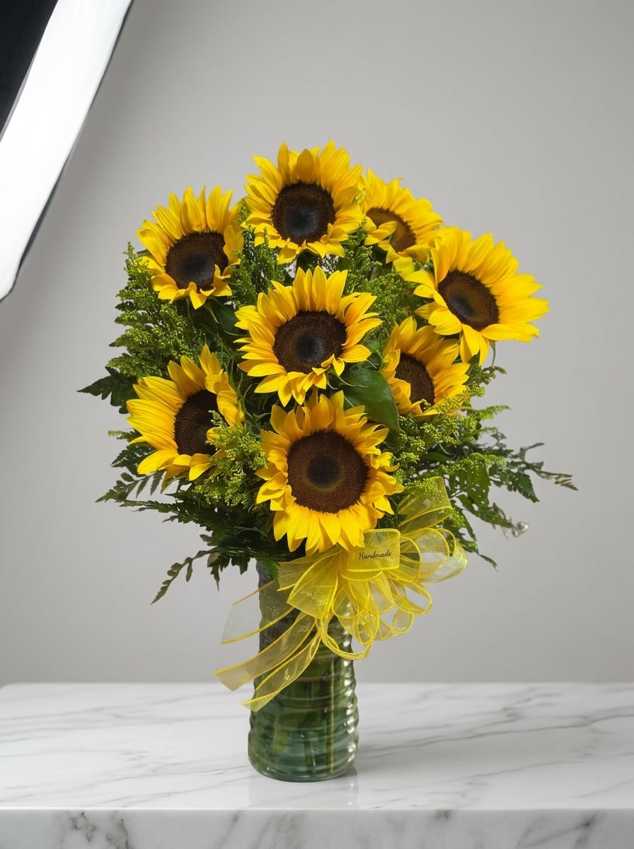 Sunflowers Vase - A cluster of vivid yellow sunflowers with bold dark centers creates an instant mood lift - perfect as a birthday bouquet, a cheerful get-well gift, or a bright Housewarming arrangement for Labor Day and all your autumn or summer celebrations. The sunflower arrangement reads as joyful and confident; the person you send it to will feel noticed and genuinely cheered by its sunny presence.  Tightly gathered for a full, rounded shape, these sunflowers are set in a clear glass vase and finished with a sheer yellow ribbon bow for a polished, gift-ready look. Lush green filler and foliage add texture and contrast, making the yellow pop while keeping the design fresh and natural. Ideal for occasions from Thanksgiving, Graduation, and Anniversary to Just Because, Thank You, or Thinking of You - this easygoing bouquet also suits weddings, prom tables, new baby visits, and holiday displays. Vase dimensions and care instructions are included at checkout so the recipient can enjoy long-lasting blooms.