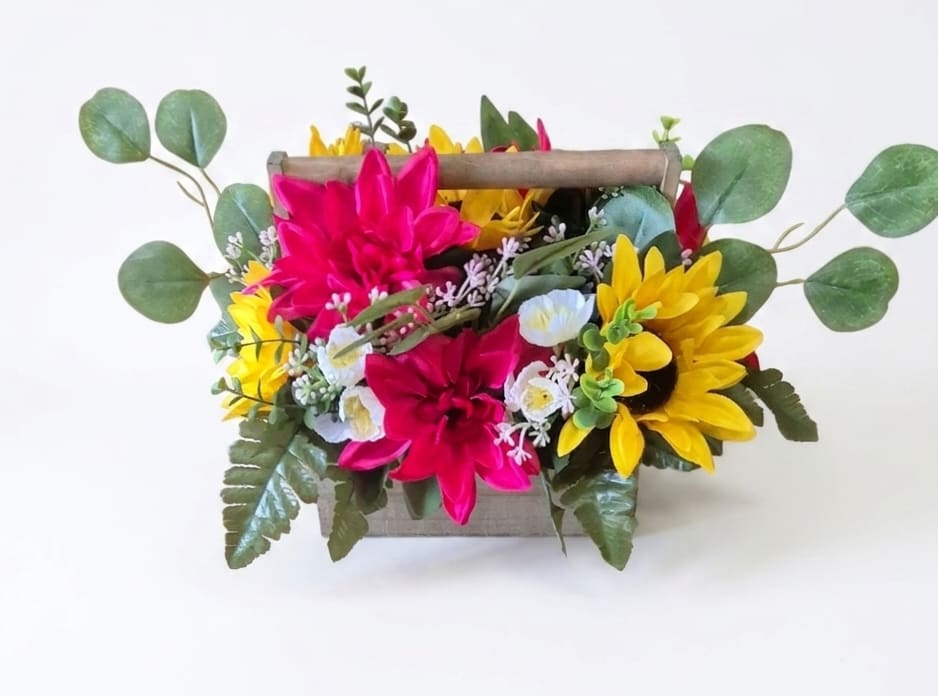 Silk Sunny Sunflower Meadow - Silk hot pink gerber blooms and sunny yellow sunflower's pop from a petite wooden container with a handle, making this casual flower arrangement a joyful pick for a birthday bouquet, springtime gift, or just-because surprise. The bright, playful color palette and garden-inspired silhouette say "cheer up" or "thinking of you" in a way that feels effortless and sincere - the kind of gift that will make the person you're thinking of smile the moment they see it.  Silk flowers are arranged tightly for a full, upbeat look, with soft, round eucalyptus-like sprigs extending outward to soften the edges and add natural movement. Tiny white filler blooms are tucked between larger flowers for depth and a layered, hand-gathered feel. The arrangement sits in a small wooden container with a handle, making it easy to display on a table or counter; its compact size is perfect for dorm rooms, office desks, or as a cheerful accent in smaller spaces. 12" Long 9" Tall 