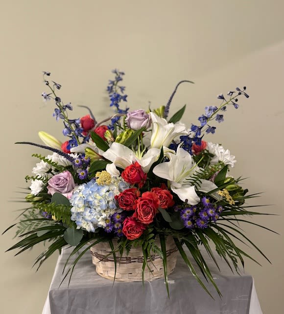 Petal Poetry - A large, garden-style flower arrangement that feels like a poem in petals-perfect as a birthday basket, anniversary surprise, sending your condolences or a  thoughtful thank-you gift. This flower arrangement makes the recipient feel celebrated and seen: the tall blue accents rise like graceful punctuation above a full cascade of blooms, while the mix of colors - white, pink, purple, blue with lively red accents - reads as joyful yet refined.  The woven basket holds an abundant, layered composition of white lilies, roses in red and lavender, a pale blue hydrangea cluster, daisies, and other seasonal blooms. Rounded blossoms sit alongside airy spikes for contrast and texture, giving the piece both fullness and movement. It's a generous size with a strong focal height from the tall blue flowers and a broad, garden-like spread across the basket.  Please note:  To ensure the freshest product there may be substitutions. 