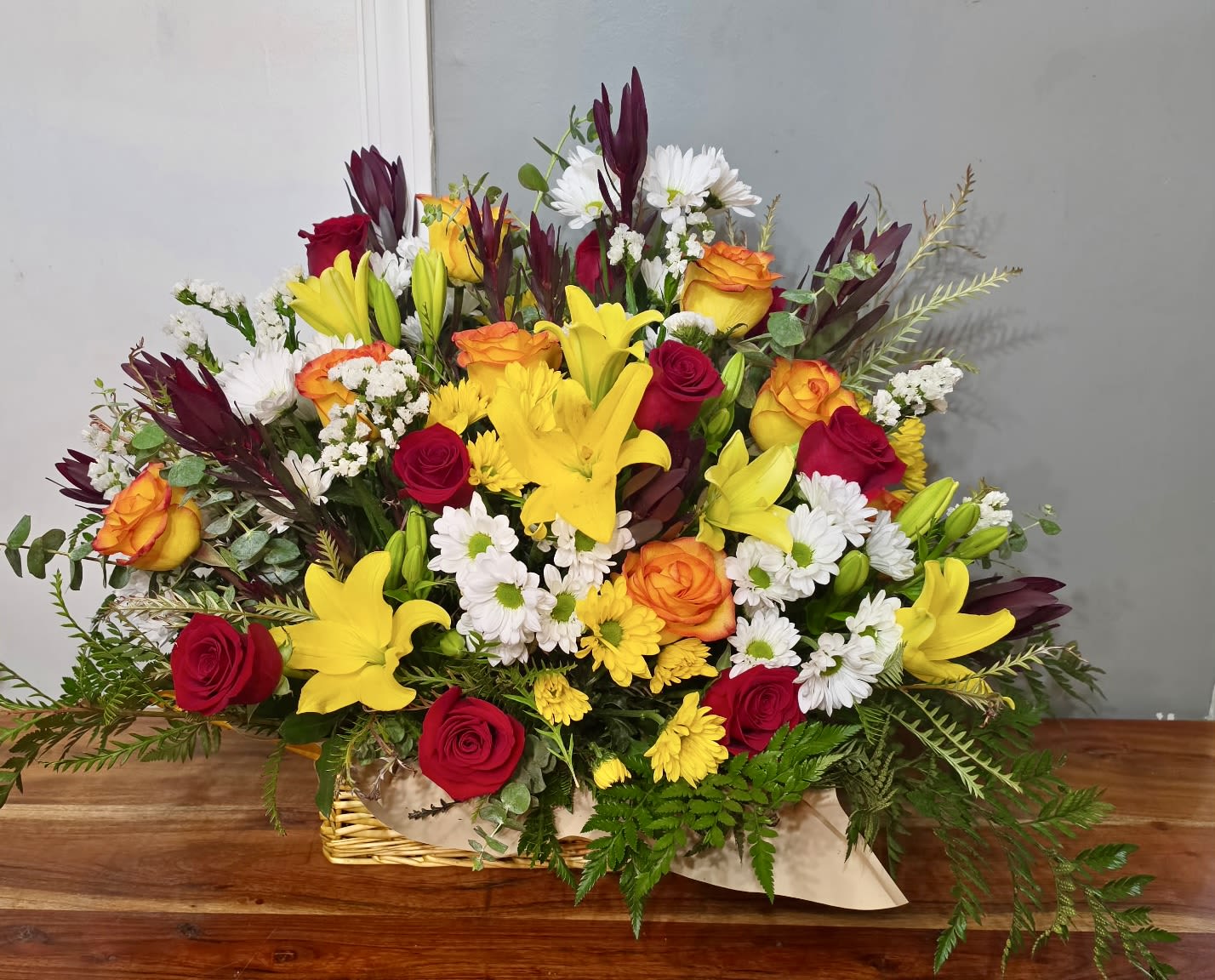 Autumn Sunshine - Bright yellow lilies and vibrant red and orange roses burst from a low, hand-wrapped basket in this Autumn Sunshine flower arrangement-perfect as a birthday bouquet, a congratulations gift, or a joyful pick-me-up for someone special. The eye-catching mix of warm fall tones and crisp white daisy-like blooms delivers an uplifting, celebratory impression that will make the recipient feel noticed and cheered.  This large, full mixed arrangement is densely packed into a low basket with the stems gathered and tied inside a rustic kraft paper wrap for a hand-delivered, abundant presentation. Expect showy lilies, classic roses in red and orange, clusters of white daisies and chrysanthemums, plus complementary filler florals that add texture and depth. The combination creates strong color contrast and a polished, slightly formal silhouette that still feels warm and approachable.  A thoughtful choice for birthdays, congratulations, housewarmings, or "just because" moments, Autumn Sunshine reads as both festive and sincere-the kind of arrangement that brightens a room and makes the person you're thinking of feel truly celebrated. Arrives sized as a generous centerpiece-style bouquet; trim stems and refresh water daily to keep blooms looking their best.