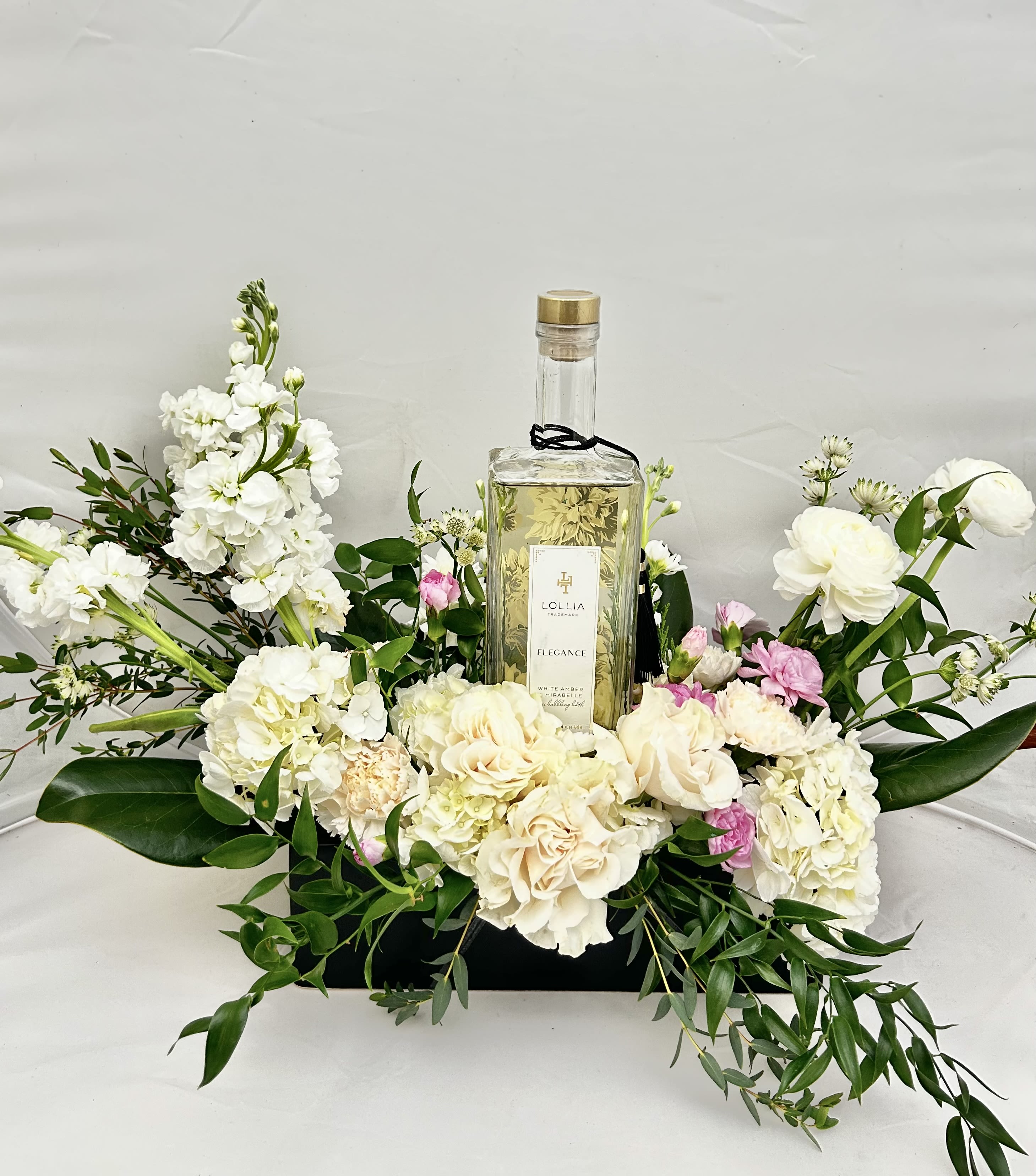 White Amber Elegance - Experience a moment of pure serenity with this sophisticated pairing. This arrangement features a lush, low-profile design of crisp white hydrangeas, delicate roses, and airy stock, accented by soft touches of pink and fresh greenery. Nestled at the heart of the blooms is a bottle of Lollia’s Relax Bubble Bath (White Amber &amp; Mirabelle), making it the perfect gift for someone who deserves a luxurious escape.