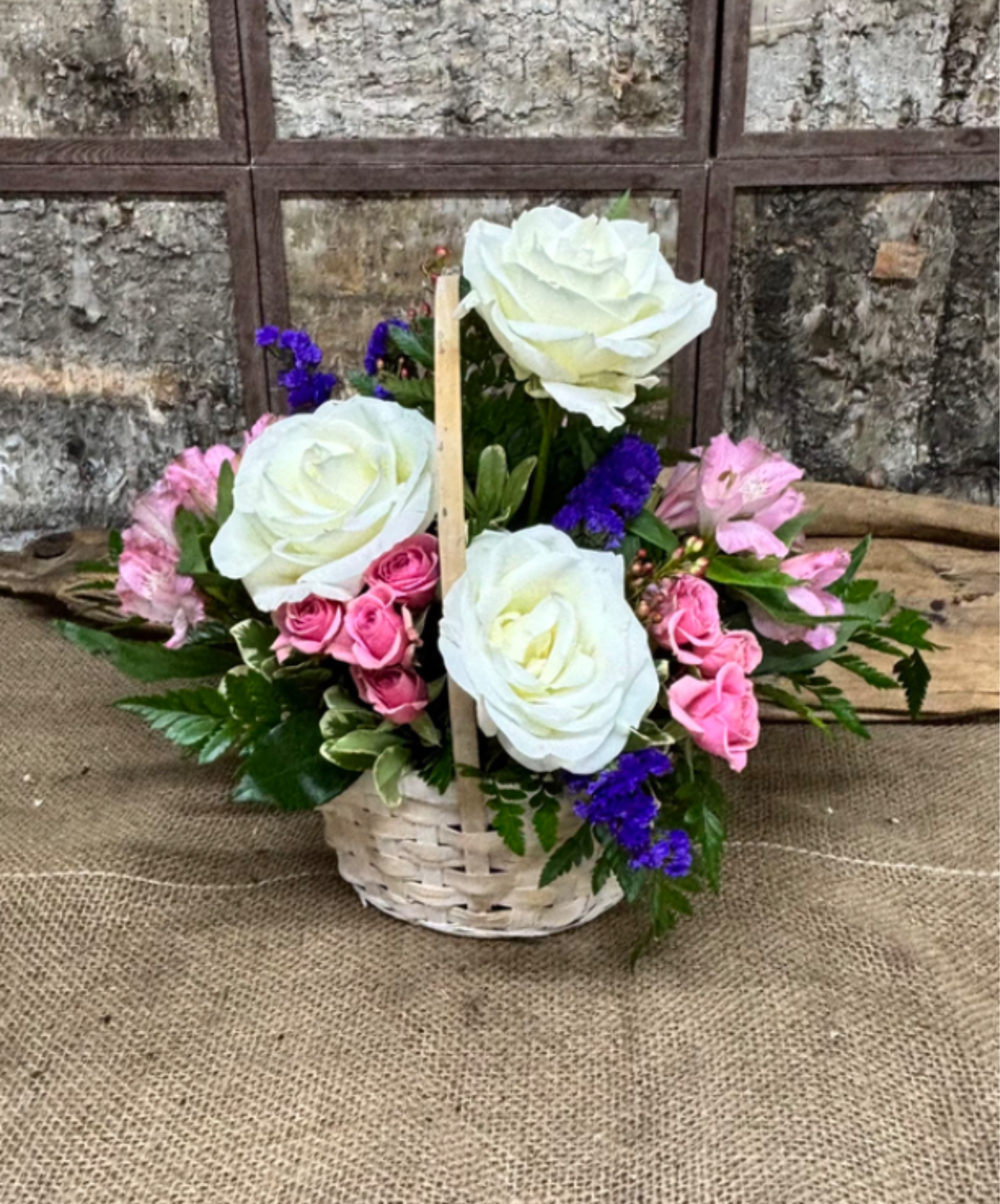 Porch Light Basket - Three large white roses form the luminous centerpiece of this Porch Light Basket, a romantic flower arrangement perfect for birthdays, anniversaries, or a thoughtful thank-you. Soft pink spray roses and tiny purple filler flowers curl around the focal blooms so the person you're gifting will feel genuinely celebrated by the gentle, uplifting color story.  Arranged in a white woven basket with a vertical wooden handle and wooden pick, the handcrafted, rustic presentation feels homey on a porch table or entryway. Compact and rounded, the mix of creamy petals, green foliage, and lively purple accents makes this an easy everyday gift or a sweet way to mark a special moment-display as shown and keep stems in fresh water away from direct heat to extend bloom life.