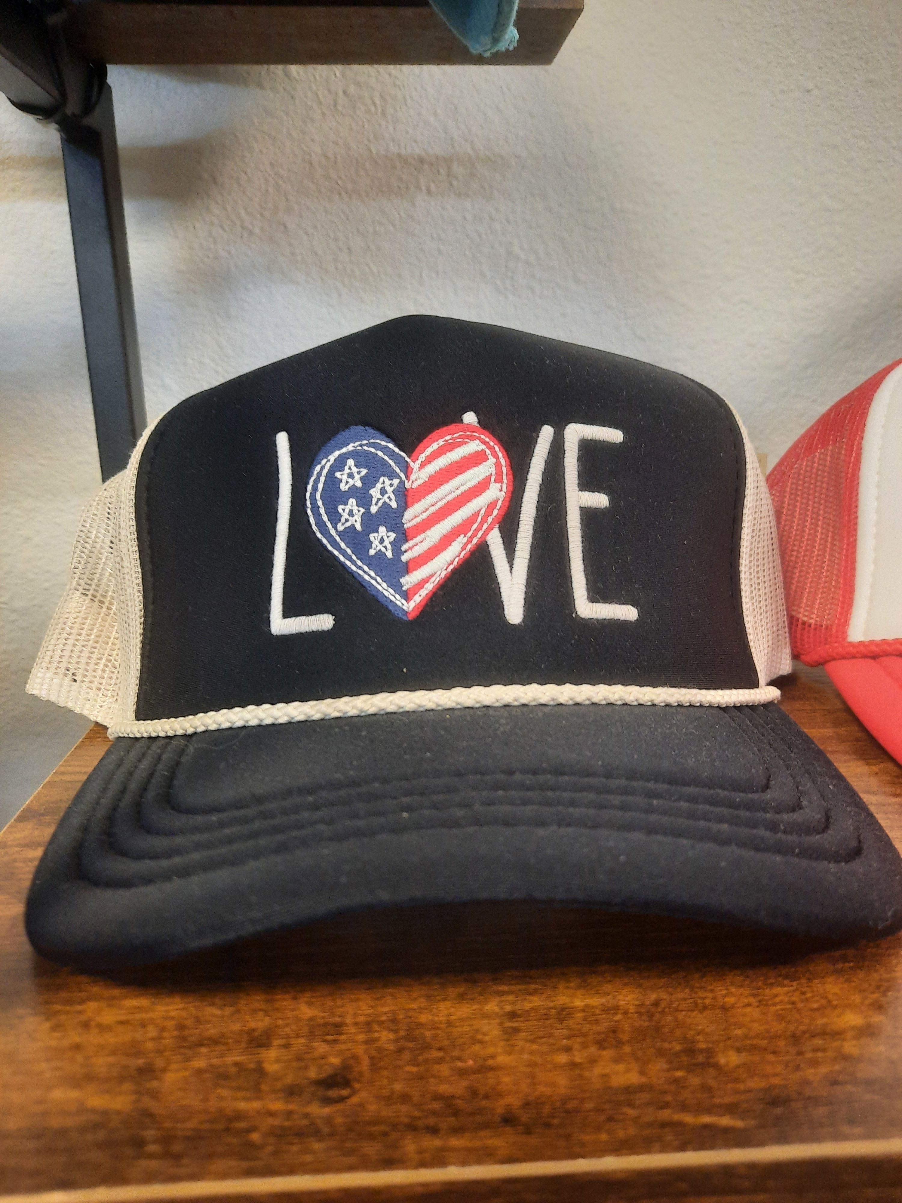 Love Trucker Hat - A playful twist on a gift-ready accessory, this Love trucker hat is a casual keepsake perfect for a summer celebration or a thoughtful "just because" gift. The black trucker-style cap reads LOVE across the front-its O replaced by a split red, white, and blue heart with star details-making it a spirited choice for Independence Day, a patriotic birthday, or to cheer on someone who loves Americana style.  Crafted with a curved brim and white mesh side panels, the hat pairs graphic embroidered lettering with a handmade, slightly vintage feel that will make the recipient smile. The contrast of black fabric, crisp white embroidery, and the tri-color heart gives it bold visual impact while staying wearable and relaxed.  Give this as a casual token to someone who appreciates novelty accessories-a friend heading to a parade, a parent to wear at a family cookout, or anyone who likes playful, giftable items. Available in one standard trucker fit; spot-clean the front panel and wipe the mesh clean to keep the embroidery bright.