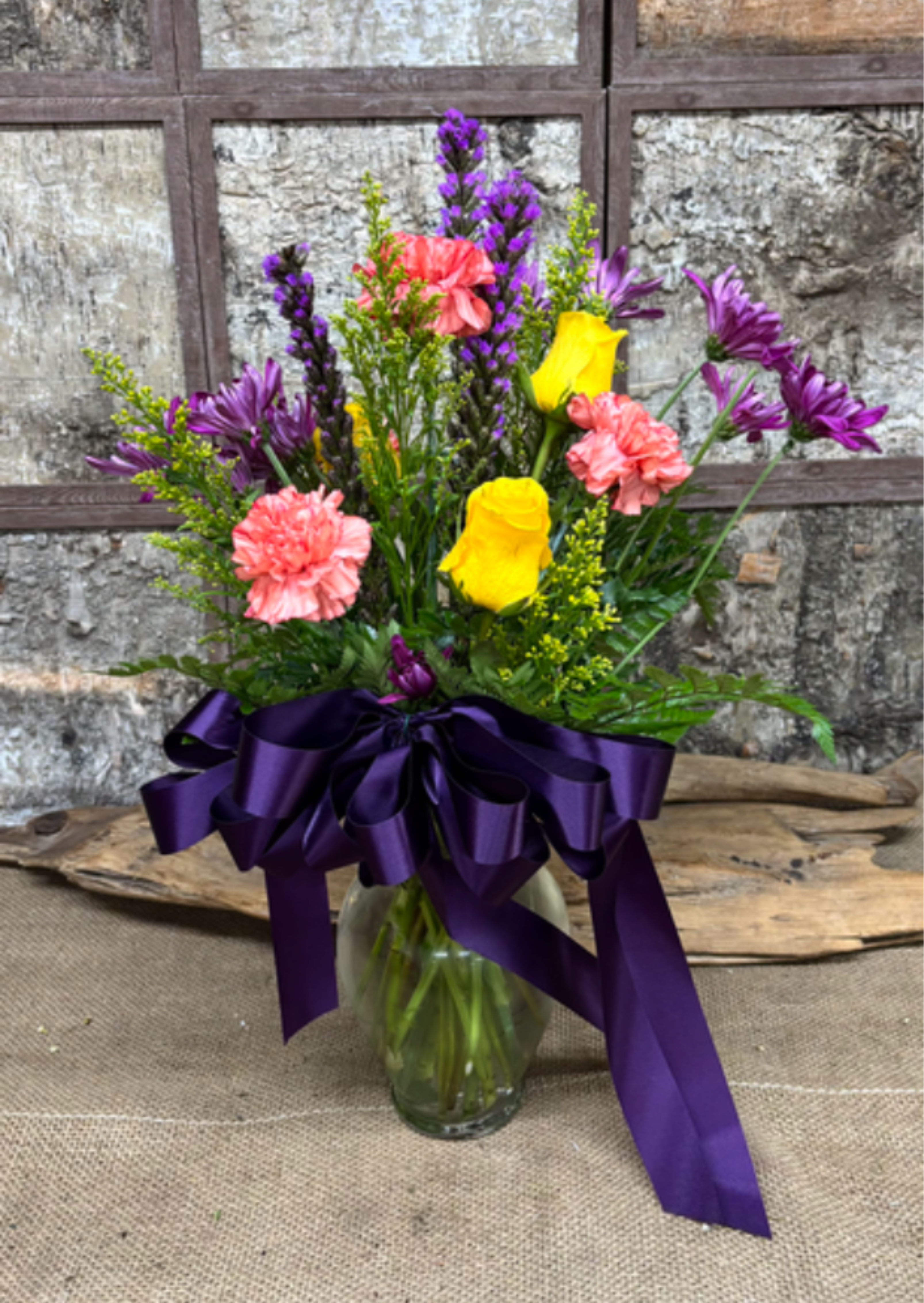 Secret Garden - Bright yellow roses and soft pink carnations mingle with purple aster-like blooms in a loose, garden-style bouquet - a cheerful flower arrangement perfect for birthdays, congratulations, thank-you gestures, or just because. The upright, airy silhouette and long stems give it a fresh-picked feel that will make the person you're thinking of smile the moment it arrives.  This bouquet is presented in a clear glass vase and finished with a large deep-purple satin ribbon for a gift-ready look; custom ribbon requests are available. Expect a lively mix of roses, carnations, asters and airy filler in purple, pink, yellow and green tones, arranged to showcase texture and color contrast without feeling fussy.  Ideal for brightening a desk, bedside table, or entryway, Secret Garden brings celebratory energy to spring occasions and everyday gifting alike. Vase dimensions and stem lengths vary with seasonal availability; for best results, trim stems and refresh the water every few days to keep blooms looking their freshest.