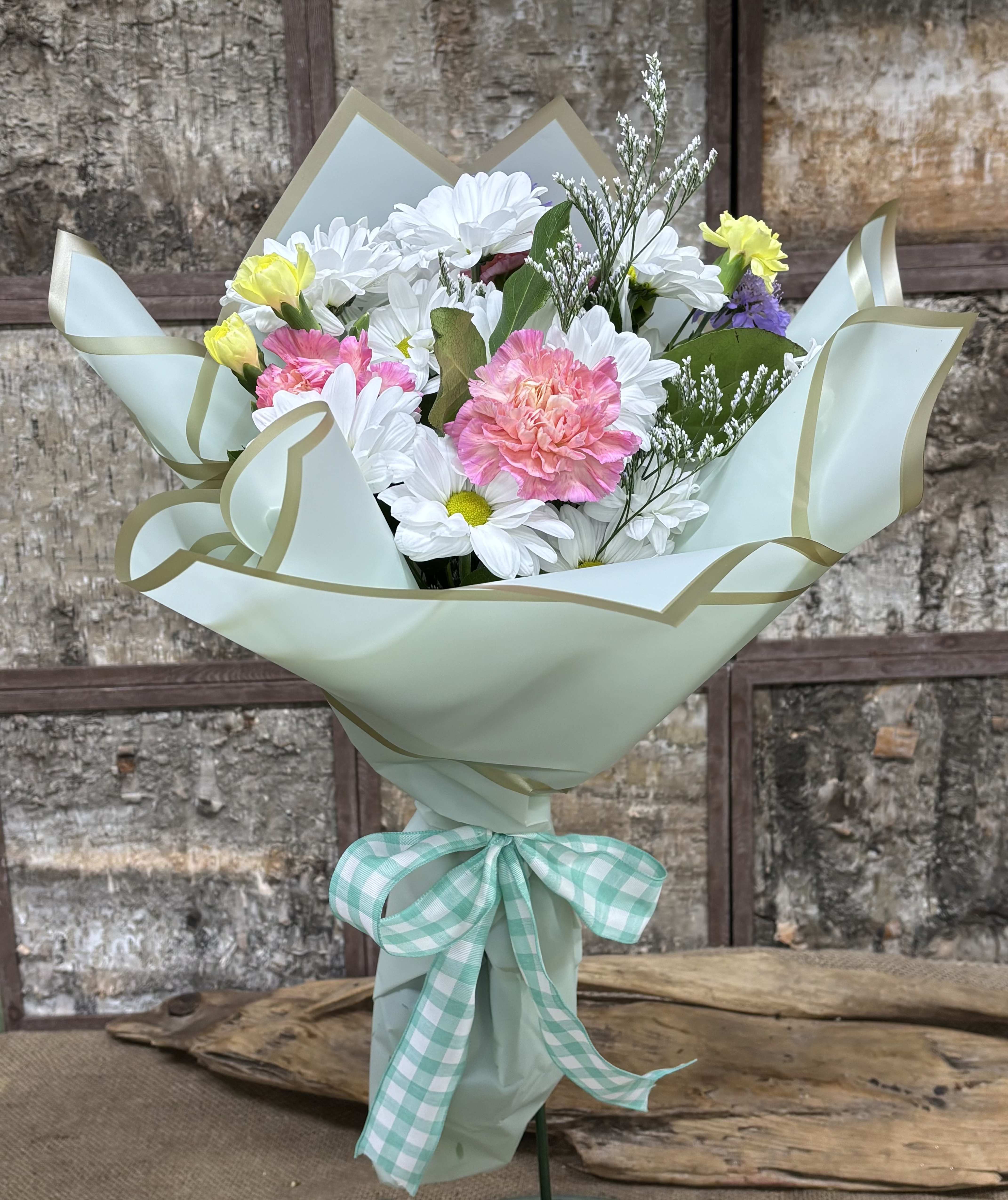 Mint Julep - Soft white daisy-like blooms paired with pink carnations give this Mint Julep flower arrangement a fresh, cheerful look - perfect as a birthday bouquet, spring celebration gift, or a thoughtful thank-you. The pale mint wrap with gold edging and mint-and-white gingham ribbon adds a handmade, charming touch that makes the recipient feel genuinely seen.  Small yellow and purple accents brighten the mix while keeping the palette pastel and gentle. Wrapped ready to give in floral paper and tied with ribbon, this everyday bouquet arrives compact and easy to display - a simple, pretty way to lift someone's day.