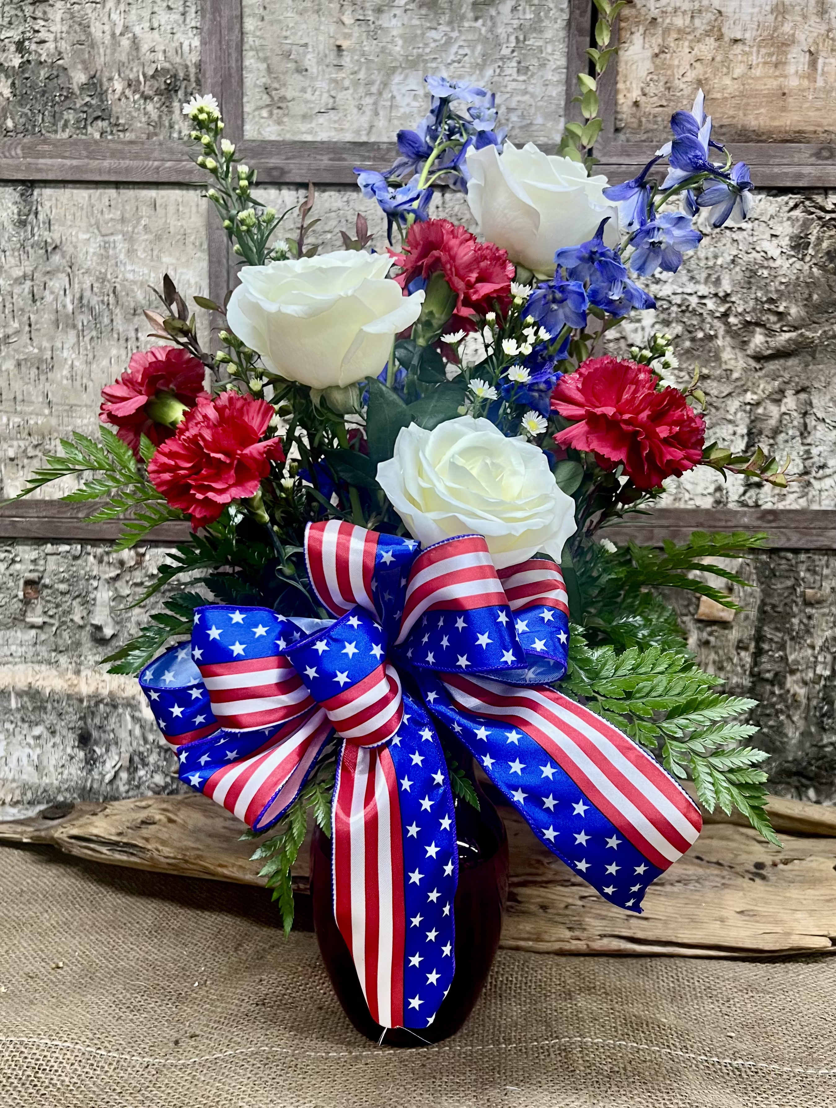 Stars &amp; Petals - A bold, patriotic statement that makes the moment feel like a celebration-this Stars &amp; Petals flower arrangement is perfect for summer holidays and national commemorations. The upright, full shape reads festive at first glance, so it's an ideal Fourth of July, Memorial Day, or Veterans Day centerpiece, or a thoughtful everyday gift to honor someone who loves red, white, and blue.  White roses mingle with bright red carnations and tall blue delphinium, while small white daisy-like fillers add a cheerful, airy touch. The bouquet is gathered in a dark glass vase and finished with a large red, white, and blue ribbon bow at the front, giving the display strong color contrast and a polished, ready-to-present look.  Send this to make the person you're thinking of feel recognized and celebrated-whether it's to thank a veteran, deck a picnic table, or brighten a patriotic gathering. The vase makes arranging easy on arrival; keep blooms in fresh water, recut stems occasionally, and enjoy peak color through the holiday weekend.