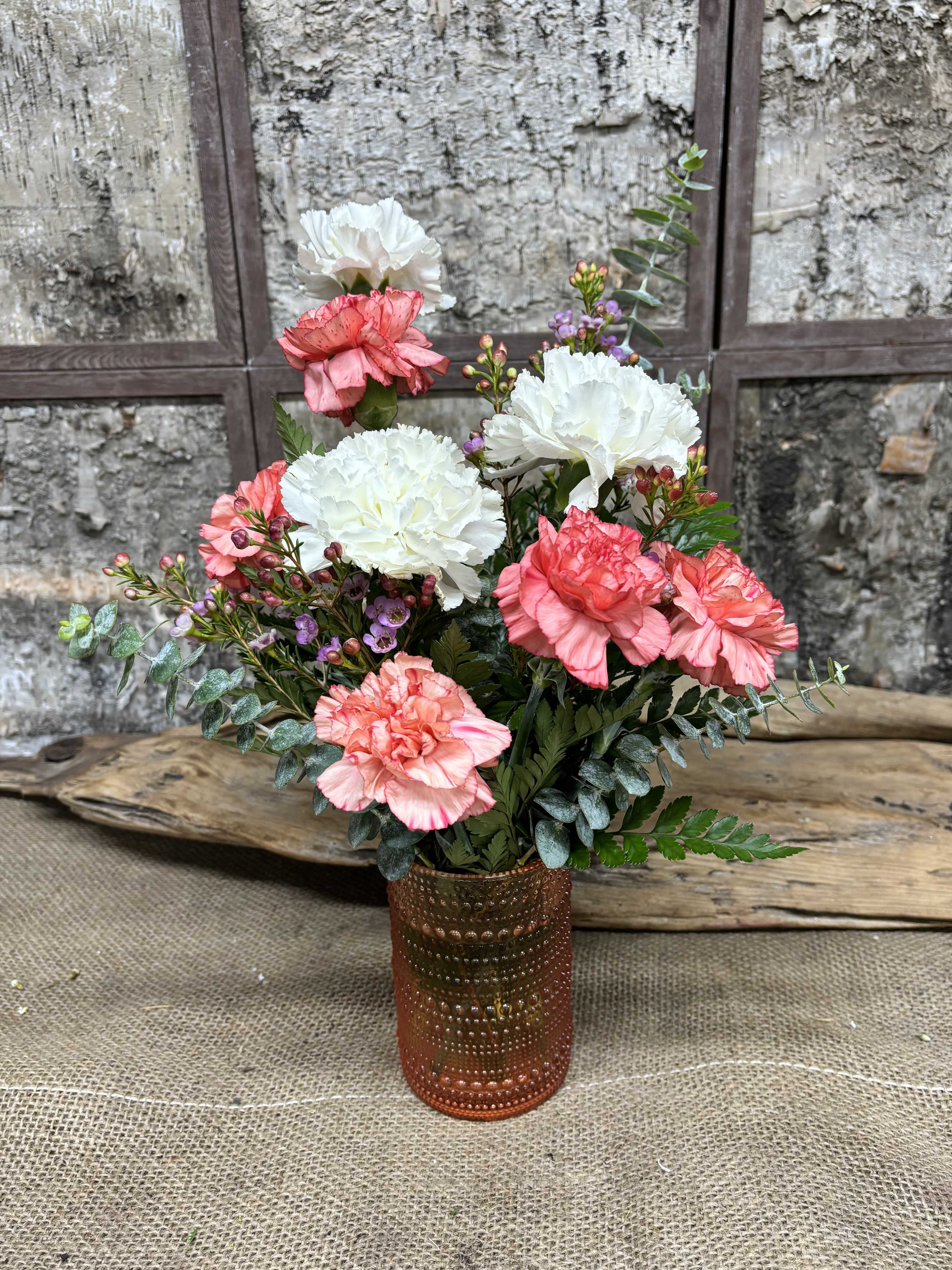 Coral Kiss - Ruffled pink and white carnations make an immediate, cheerful impression in this Coral Kiss flower arrangement - an ideal birthday bouquet, thank-you gift, or springtime surprise. The compact upright design keeps the focus on the soft, romantic blooms so the person you're thinking of will feel warmly noticed the moment they see it.  Small purple filler blooms and silvery-green eucalyptus-like foliage add delicate texture and contrast around the larger carnations, giving the arrangement a layered, garden-fresh look. The flowers are presented in a copper-toned textured vase that lends a rustic, handcrafted warmth and looks beautiful on a mantel, bedside table, or kitchen counter.  Designed for everyday gifting, Coral Kiss suits friends, coworkers, or loved ones who appreciate gentle color and simple elegance. Vase is included; for best longevity refresh water every 2-3 days and trim stems slightly when re-cutting for display.
