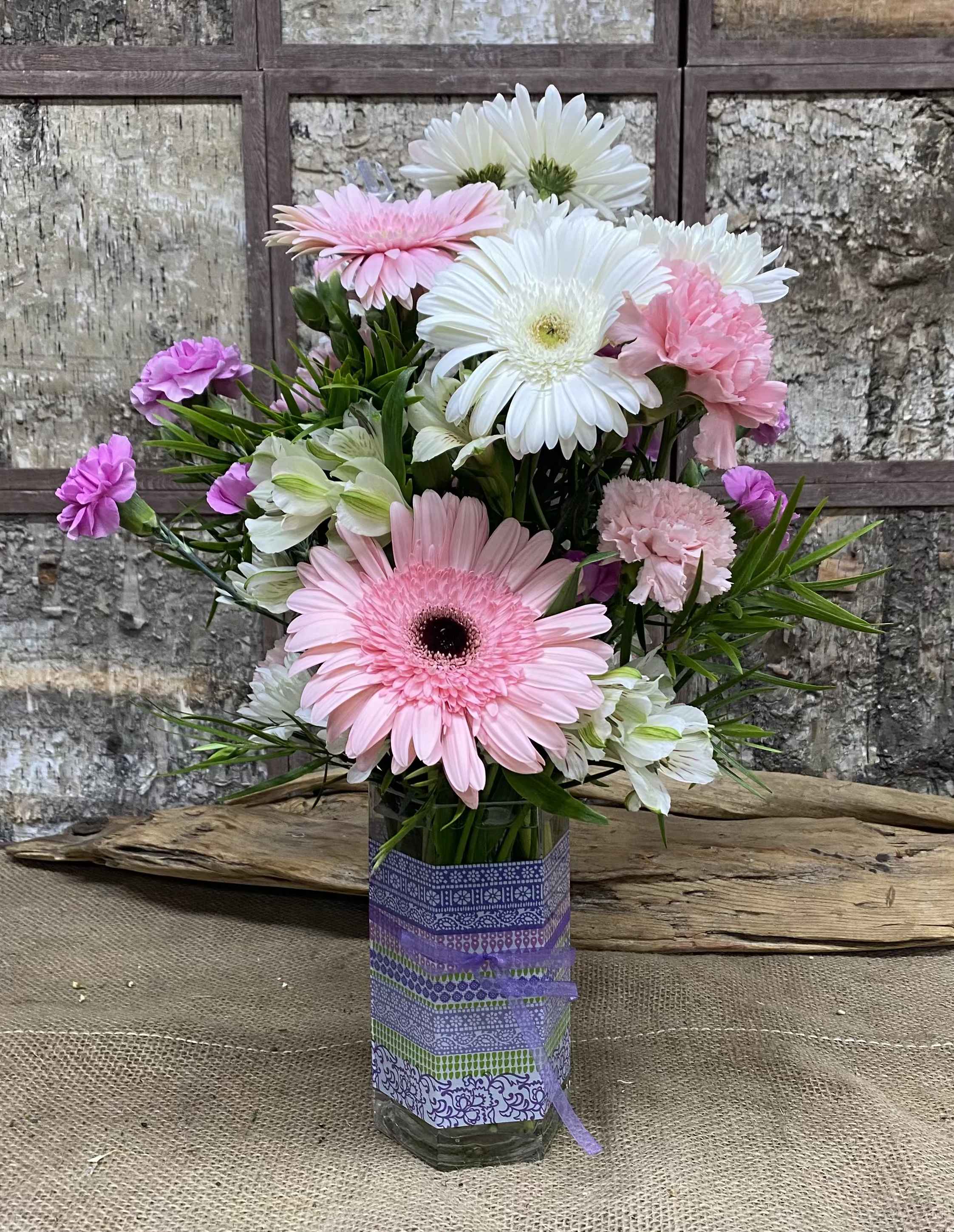 Blushing Skies - Soft pink and lavender blooms create an uplifting, garden-fresh impression - perfect for a birthday bouquet, thank-you gift, or simple springtime surprise. This flower arrangement blends gerbera daisies, carnations, and alstroemeria to deliver a cheerful, everyday bouquet that will make the person you're thinking of feel noticed and celebrated.  The arrangement pairs pink and white gerbera daisies with pale pink and lavender carnations and touches of alstroemeria for delicate texture and contrast. Blooms are arranged at varied heights for a full, rounded look inside a clear glass vase wrapped in a purple patterned sleeve and tied with a decorative ribbon, giving it a handcrafted, gift-ready presentation.  Ideal for brightening a kitchen table, office desk, or bedside, this design reads casual yet thoughtful - an easy way to mark small milestones or send a warm "thinking of you." Order with same-day pickup or delivery where available; fresh-cut stems arrive ready to display in the included vase, and simple care (fresh water and a trim every few days) will keep the bouquet looking its best.