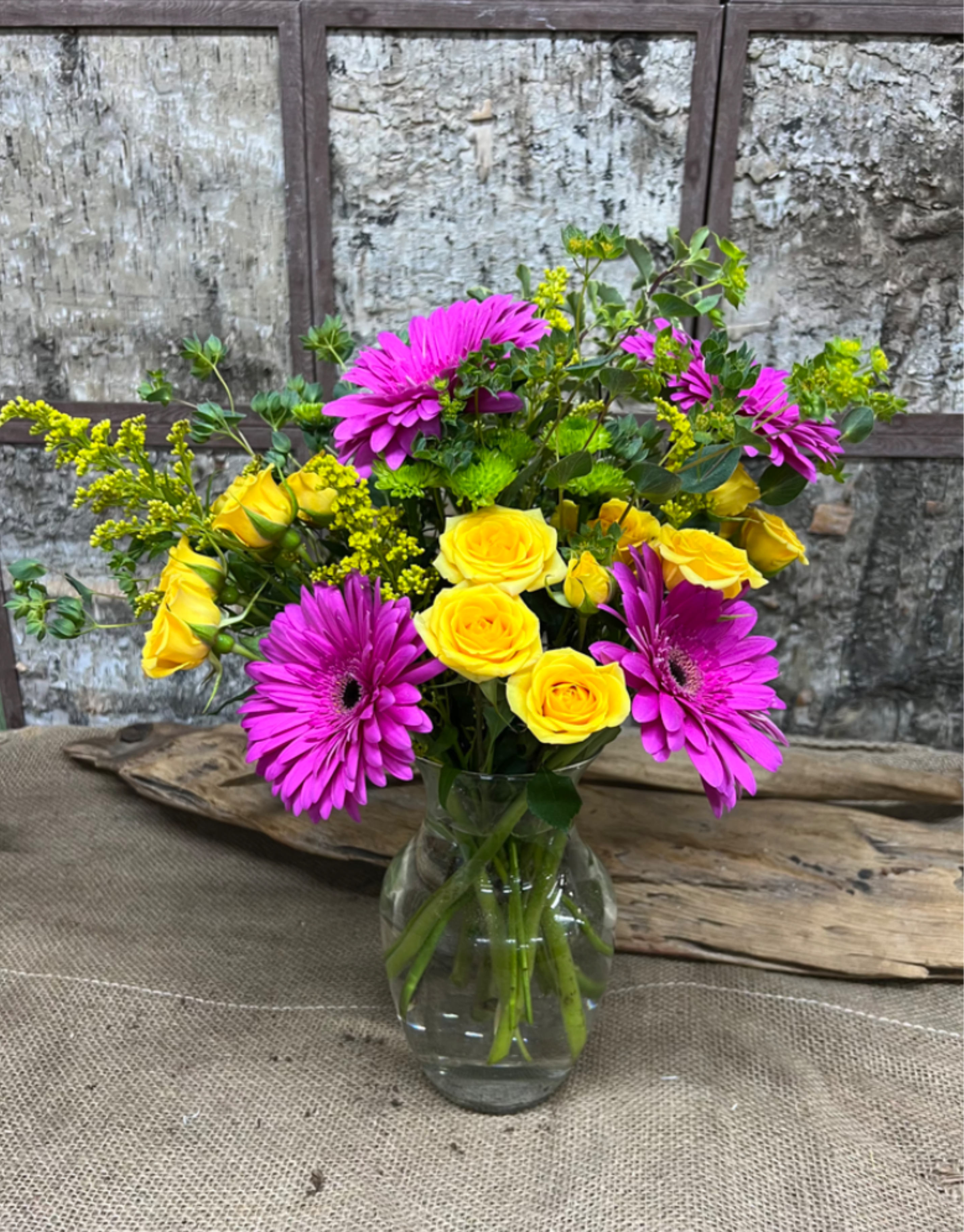 Berry Bliss - Bright yellow roses paired with vivid magenta gerbera daisies create an attention-grabbing contrast in this cheerful flower arrangement-perfect as a birthday bouquet, a springtime thank-you, or a casual "thinking of you" gift. The lively pink-and-yellow palette reads fresh and upbeat, so the person you're sending it to will feel brightened and genuinely celebrated the moment it arrives.  Set in a clear glass vase, the bouquet includes small yellow filler blooms and airy greenery that soften the bold colors and give the composition a loose, garden-like texture. The mix of roses, gerbera daisies, and tucked-in fillers makes the arrangement feel full without being fussy; its compact vase presentation suits tabletops, desks, or a kitchen counter.  Ideal for everyday celebrations and informal occasions, this arrangement delivers a burst of color that lifts the mood. Vase is included; for best longevity, refresh the water every couple of days and recut stems slightly when you change the water.