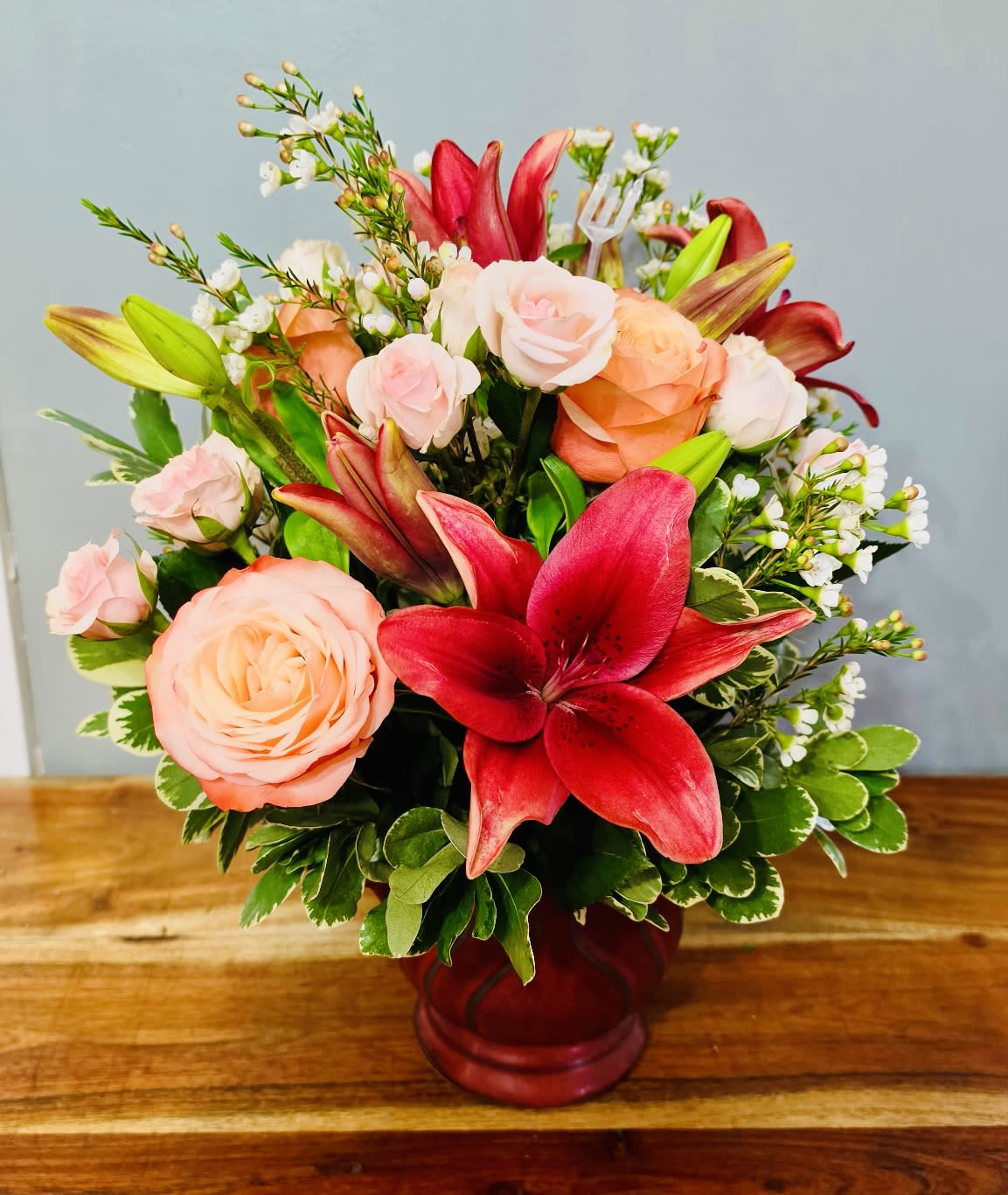 Blushing Blooms - Bold red lilies take center stage in this romantic mixed bouquet, making it an ideal anniversary roses and birthday bouquet that reads as both passionate and tender. The red glass vase amplifies the lilies' drama while a halo of blush, peach, and pale pink roses softens the composition-perfect for showing love, celebrating a milestone, or sending warm birthday wishes to someone special.  The arrangement pairs large red lilies with clusters of roses in varying pink and peach tones, accented by small white filler blossoms and variegated greenery for texture and contrast. The design is rounded and compact with a hand-tied look, creating a polished yet approachable presentation that feels lively without becoming fussy. The red glass vase ties the palette together and keeps the display low and table-friendly.  Made to make the recipient feel genuinely seen and celebrated, this bouquet works beautifully on a dinner table, bedside, or office desk. Sizes are compact and gift-ready in the included red vase; keep blooms in fresh water, recut stems when arranging, and enjoy the mix of bold color and soft fragrance for several days.