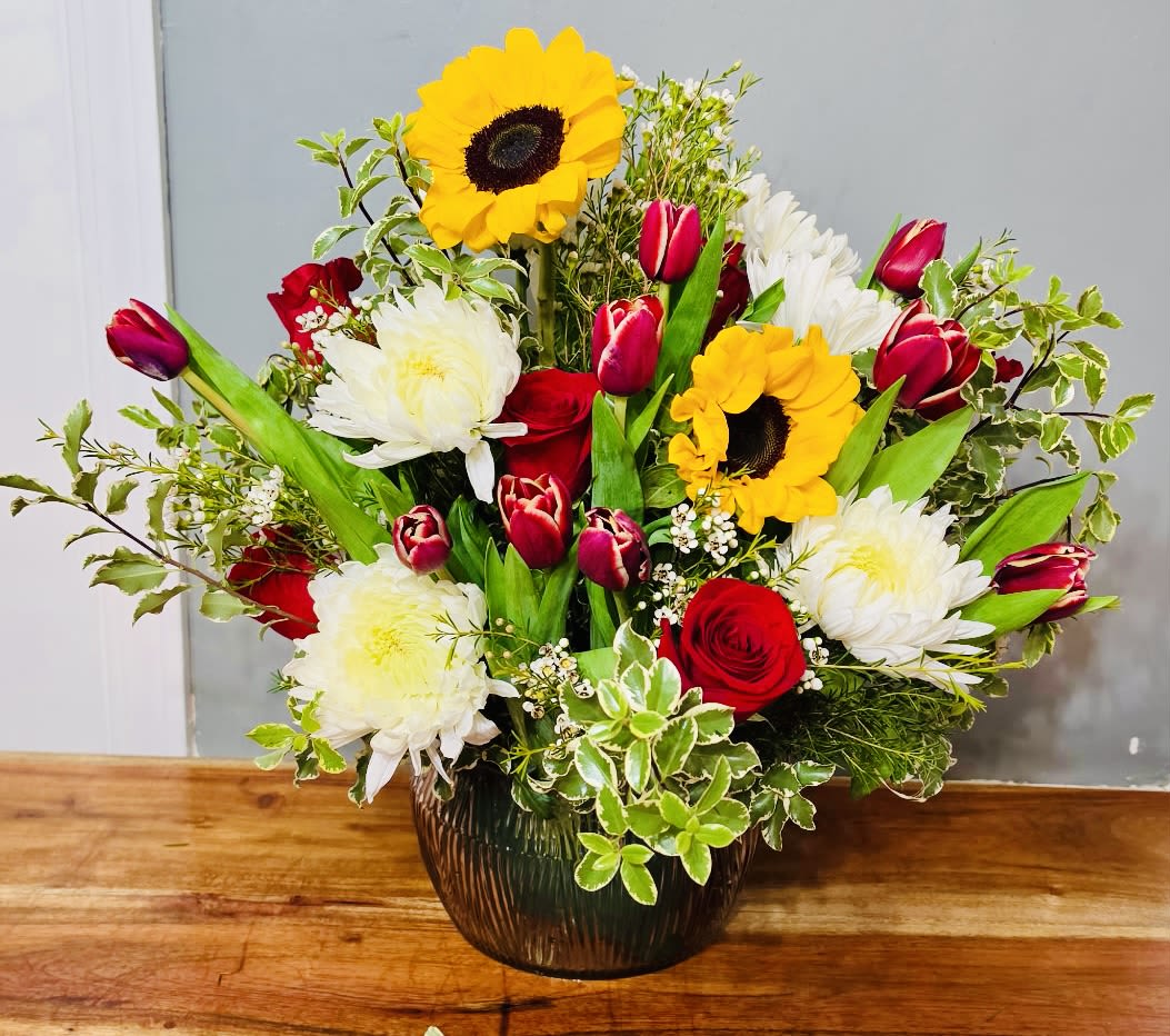 Tulip n Tango - Bright yellow sunflowers take center stage in this mixed-color bouquet, paired with red roses and red tulips for a bold, celebratory look-perfect as a birthday bouquet, congratulations arrangement, or a warm thank-you gift. The combination of sunny yellows, rich reds, and crisp whites creates an uplifting contrast that will make the person you're sending it to feel noticed and genuinely celebrated.  Arranged in a rounded dark glass vase, the design is full and layered with upright blooms rising above a dense base of mixed greenery and small white chrysanthemum-like flowers for texture and volume. The glass vase gives the bouquet a slightly formal polish while keeping the overall mood cheerful and versatile; dimensions and vase size are suited for tabletop display and easy placement in an office or home.  This is the kind of bouquet that reads joyful without being fussy-ideal for coworkers, friends, or family when you want to mark a happy moment or simply brighten someone's day. Standard care: keep water fresh and trim stems every few days to extend vase life.