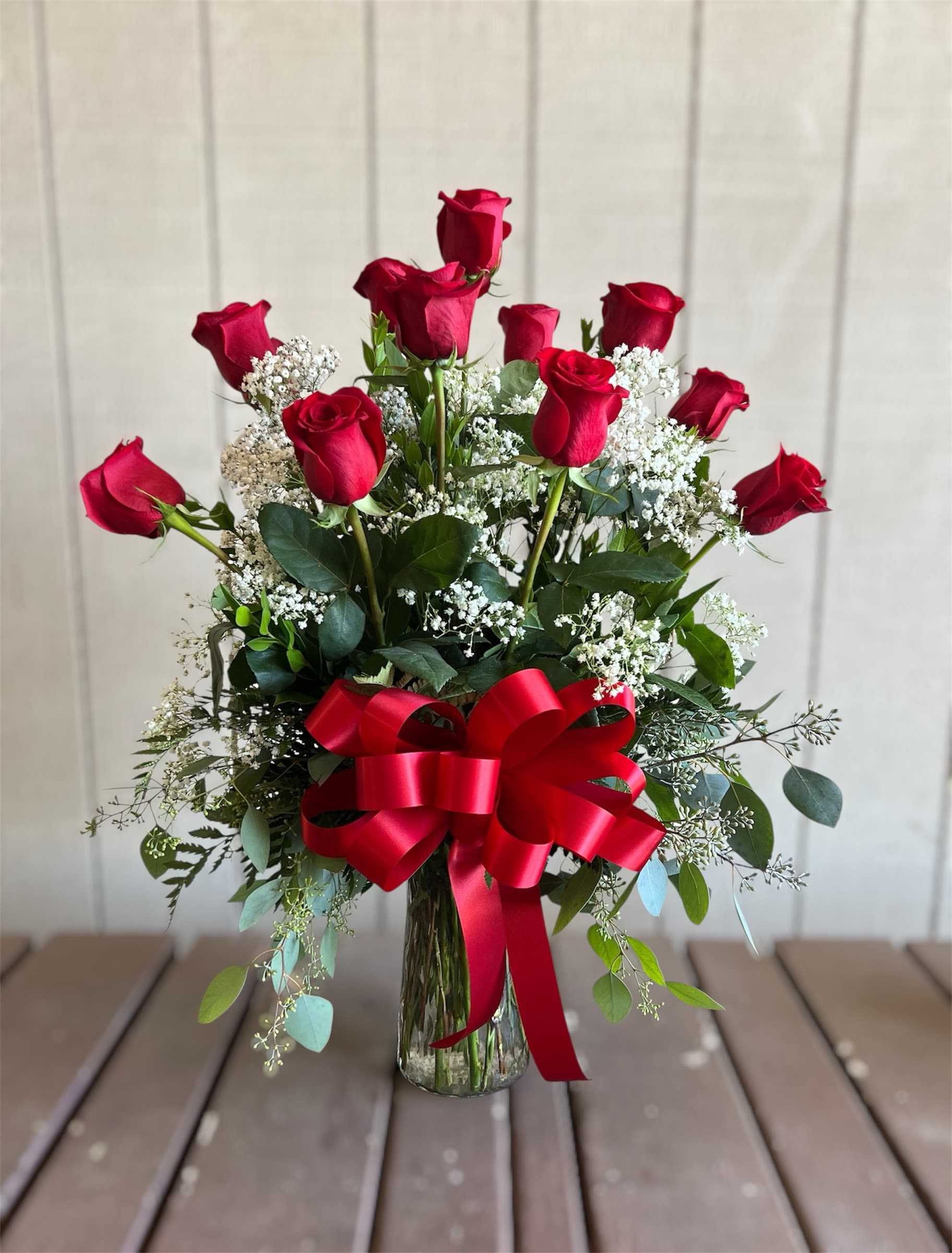 Dozen Long-Stemmed Red Roses - A classic dozen red roses arranged upright in a tall clear glass vase-this romantic bouquet makes a bold declaration for Valentine's Day, anniversaries, or any moment you want to say "I love you." Deep red blooms rise above clusters of small white filler flowers and mixed greenery, creating a striking red-white-green palette that reads as timeless and celebratory for the person you're thinking of.  The arrangement is composed of twelve long-stem red roses spaced to create height and a balanced silhouette, set in a clear glass vase and finished with a red satin bow tied around the vase for a polished, gift-ready look. Small white filler blooms and assorted greenery soften the composition and add texture, while the upright, full design ensures the bouquet reads beautifully from every angle.  Perfect for surprising a partner on a special date or marking a meaningful milestone, this bouquet sends a clear message of affection and admiration so she'll feel truly cherished. Vase-arranged and ready to display on arrival; keep the water fresh and trim stems if you transfer blooms to a different container to extend their vase life.