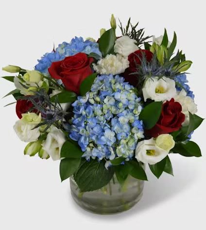 OLD GLORY - This stunning patriotic arrangement combines vibrant red roses, cool blue hydrangea, and elegant white lisianthus, with a touch of texture from the eye-catching blue thistle. It's all perfectly showcased in a clear cylinder vase, creating a gift that's as patriotic as it is gorgeous.  Whether you're hosting a summer cookout, throwing a July 4th bash, celebrating a graduation, or simply want to make a statement in your home or office, this arrangement is the perfect choice. It's a real showstopper that adds a touch of Americana to any occasion.