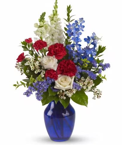 SEA TO SHINING SEA - A bold display of white roses, snapdragons and waxflower along with blue delphinium and red carnations delivered in a blue glass vase, create a powerful tribute to freedom. Approximately 14" W x 21" H