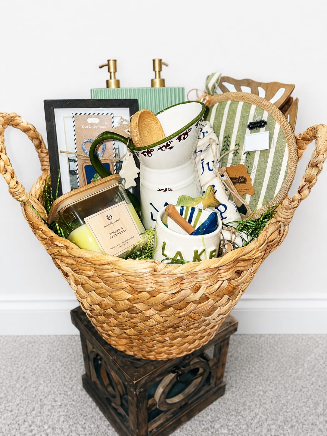 Lakehouse Theme Gift Basket - A cozy, curated Lakehouse Theme Gift Basket designed for housewarming and special gifting moments - this woven basket brings rustic charm and practical comforts to the person you're thinking of. Thoughtful as a housewarming gift set, it reads like a warm welcome: natural wicker and wood tones sit alongside soft whites, greens, blues and hints of gold to create an inviting, lived-in look that suits a kitchen, entryway, or living room.  Tucked among the arrangement are low-maintenance succulents that add a fresh green accent, plus a ceramic mug, candle jar, soap dispenser, decorative tray, picture frame, fabric pouch, wooden spoon, decorative sign and small wooden crate - everything arranged tightly for a polished presentation. Textures range from smooth ceramics and glass to woven fabric and warm wood, giving the basket both visual variety and everyday usefulness.  Perfect for new homeowners, hosts, or anyone who appreciates thoughtfully chosen home goods, this gift basket says "welcome home" without fuss. 