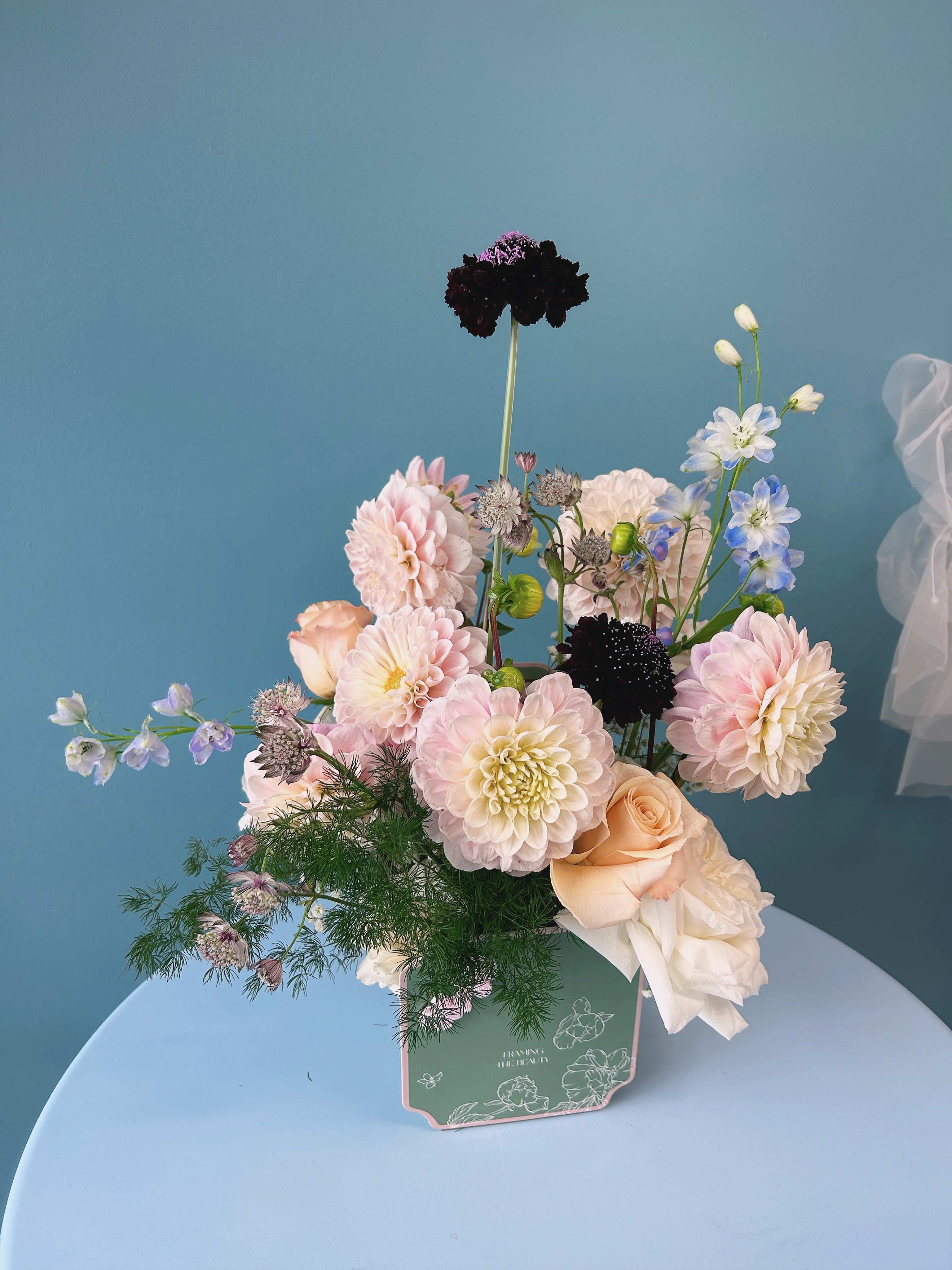 Pastel bouquet of dahlias, roses, and blue delphinium in a green box