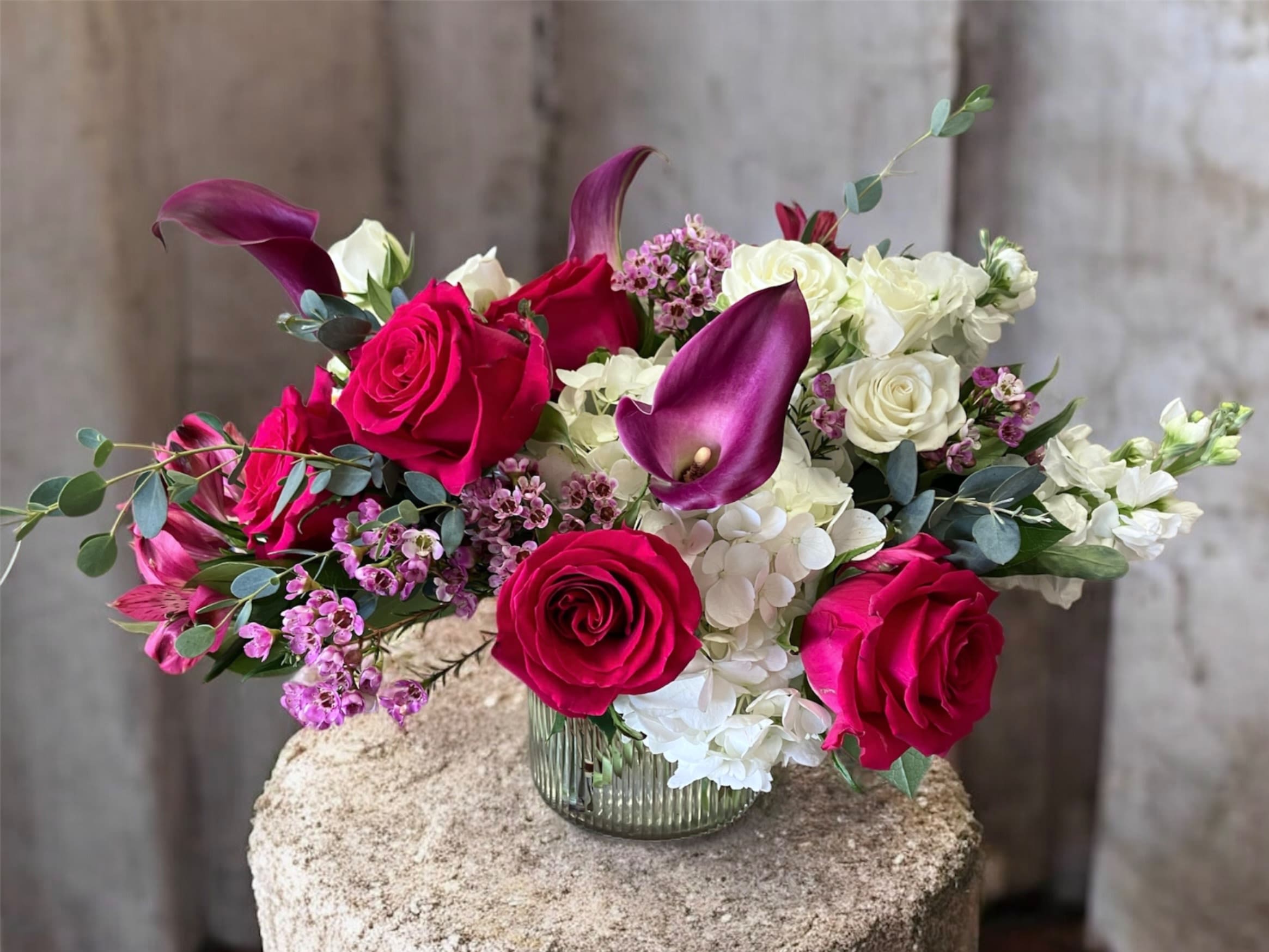 Adore You - A lush and elegant arrangement featuring soft white hydrangeas paired with vibrant hot pink roses. Accents of fragrant white stock and graceful purple calla lilies add depth and contrast, while pink alstroemeria and delicate white spray roses bring texture and movement. Finished with airy wax flower and fresh seasonal greens, this design balances bold color with timeless sophistication. Pictured as Deluxe