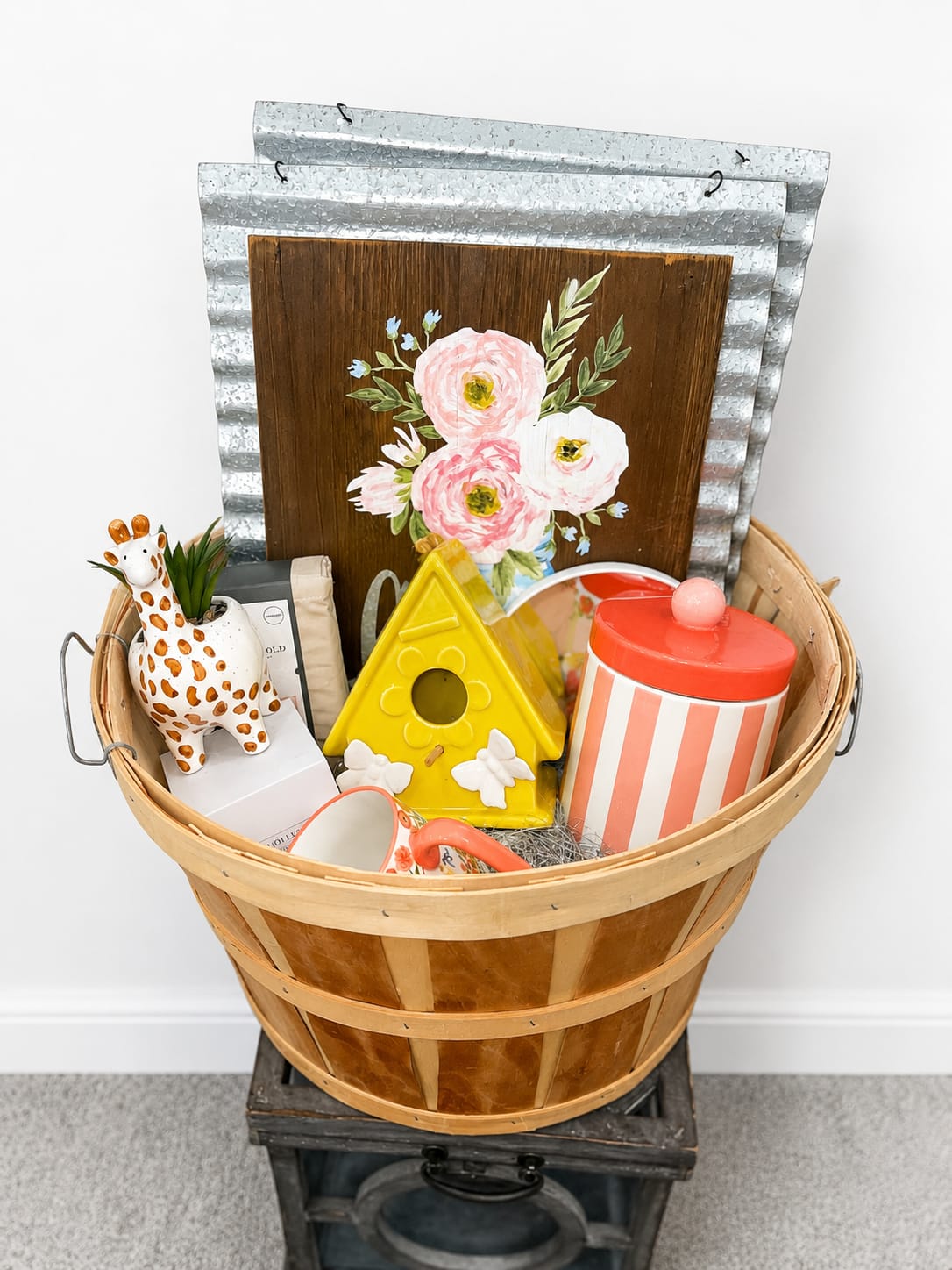 Homewarming Gift Basket  - A sunlit mix of handcrafted charm and cheerful color makes this Homewarming Gift Basket a joyful choice for welcoming someone to a new home or surprising them with a thoughtful gift set. This decorative gift basket-perfect as a housewarming present or an everyday "thinking of you"-delivers an uplifting first impression with warm wood tones and pops of pink, yellow, coral, blue and white that feel friendly and fresh.  Inside the wooden basket with metal handles you'll find a painted floral wooden sign, a whimsical ceramic giraffe planter, a bright yellow ceramic birdhouse, a striped lidded jar, a small mug, a packaged treat, a mirror and metal wall decor-an eclectic mix of shapes and textures that layers playful ceramics with useful home accents. The pieces work together to style a tabletop, shelf, or entryway; the mix of colors and handcrafted finishes gives the set an artisanal, collected-over-time look.  Give this basket to new homeowners, friends moving into an apartment, or anyone who appreciates quirky, cheerful décor-the arrangement sends a warm, celebratory message that the person you're thinking of is welcome and cared for. Items arrive arranged in the wooden basket for easy gifting; if you need delivery or timing for a housewarming celebration, select your preferred date at checkout.