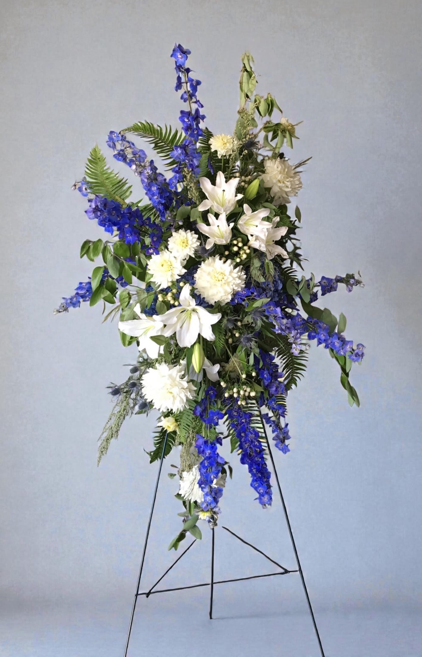 Blue Skies Easel Spray - Airy blue delphinium and white lilies arranged on an easel offering a calm and graceful memorial tribute.
