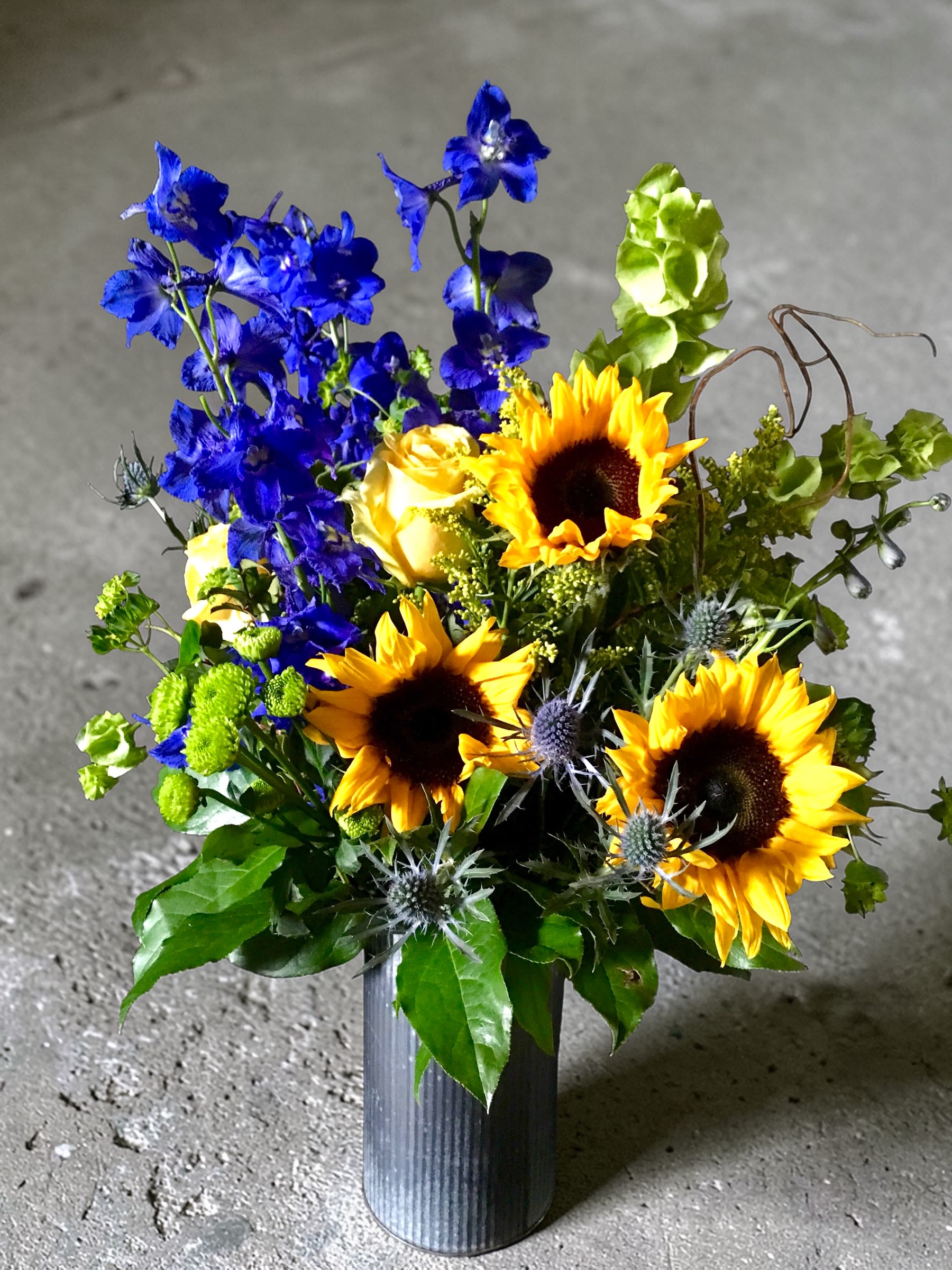 Breathe - Bright sunflowers, vivid blue delphinium, yellow roses and green fillers - a cheerful, garden-style bouquet perfect for celebrations or thank-you gifts.