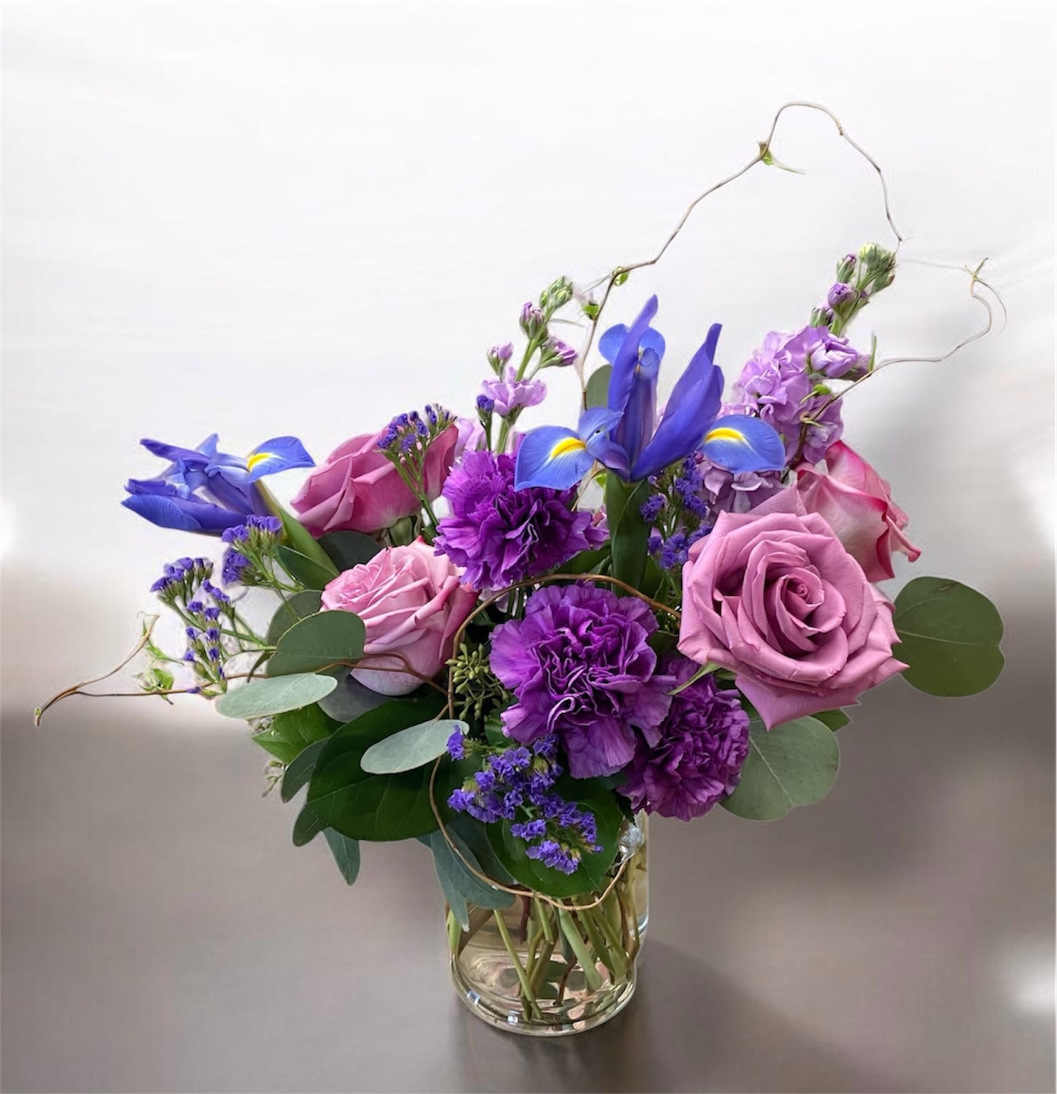 Charmer - Lavender roses, purple carnations and vivid blue iris in a clear glass vase with eucalyptus create a soothing arrangement for birthdays, Mother's Day, or just because.