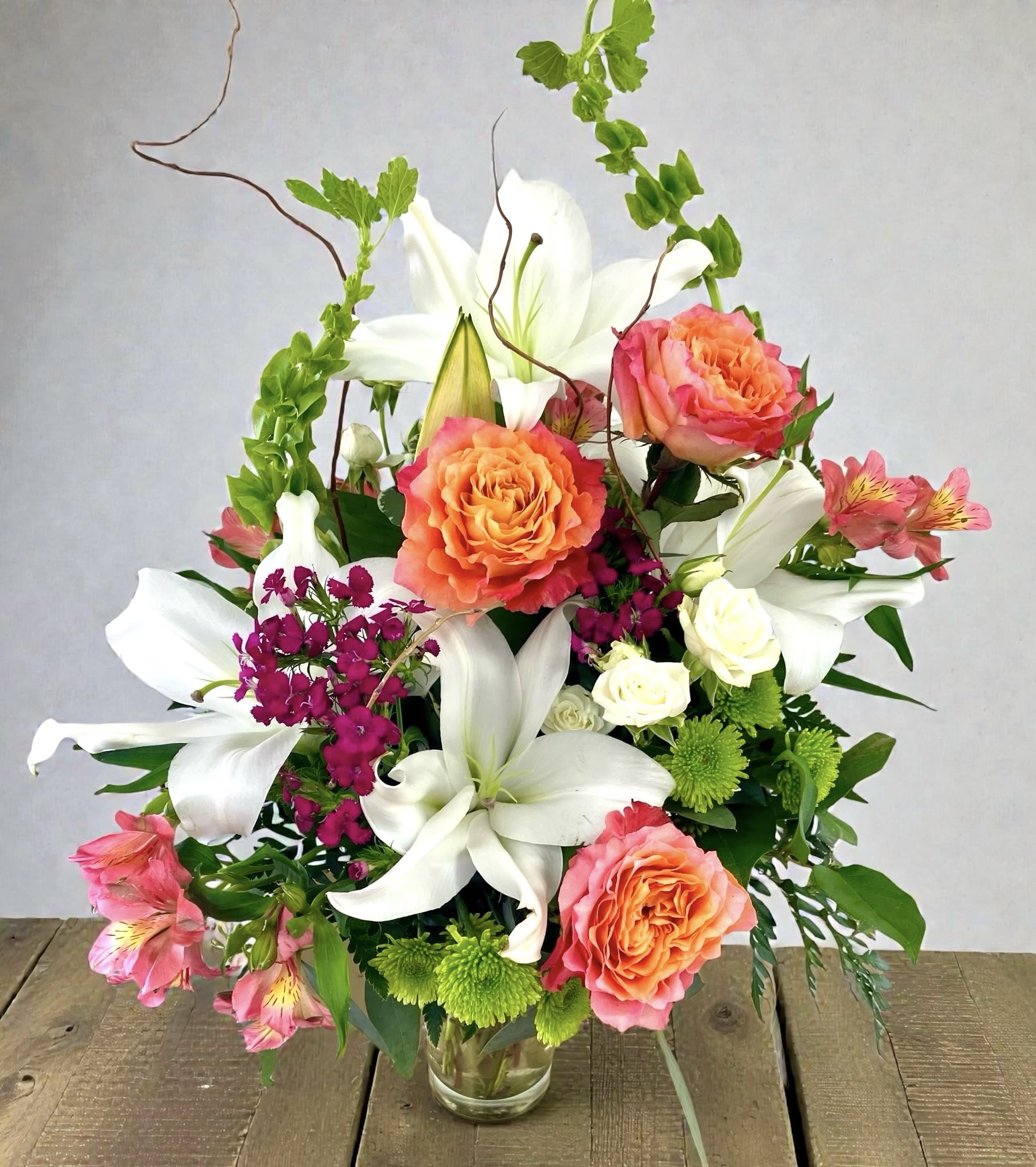 Cheeky - White lilies, coral-orange roses, pink alstroemeria, dianthus with green button mums in a clear glass vase create a fresh and lively arrangement for birthdays, thank-yous, or sympathy for the home.