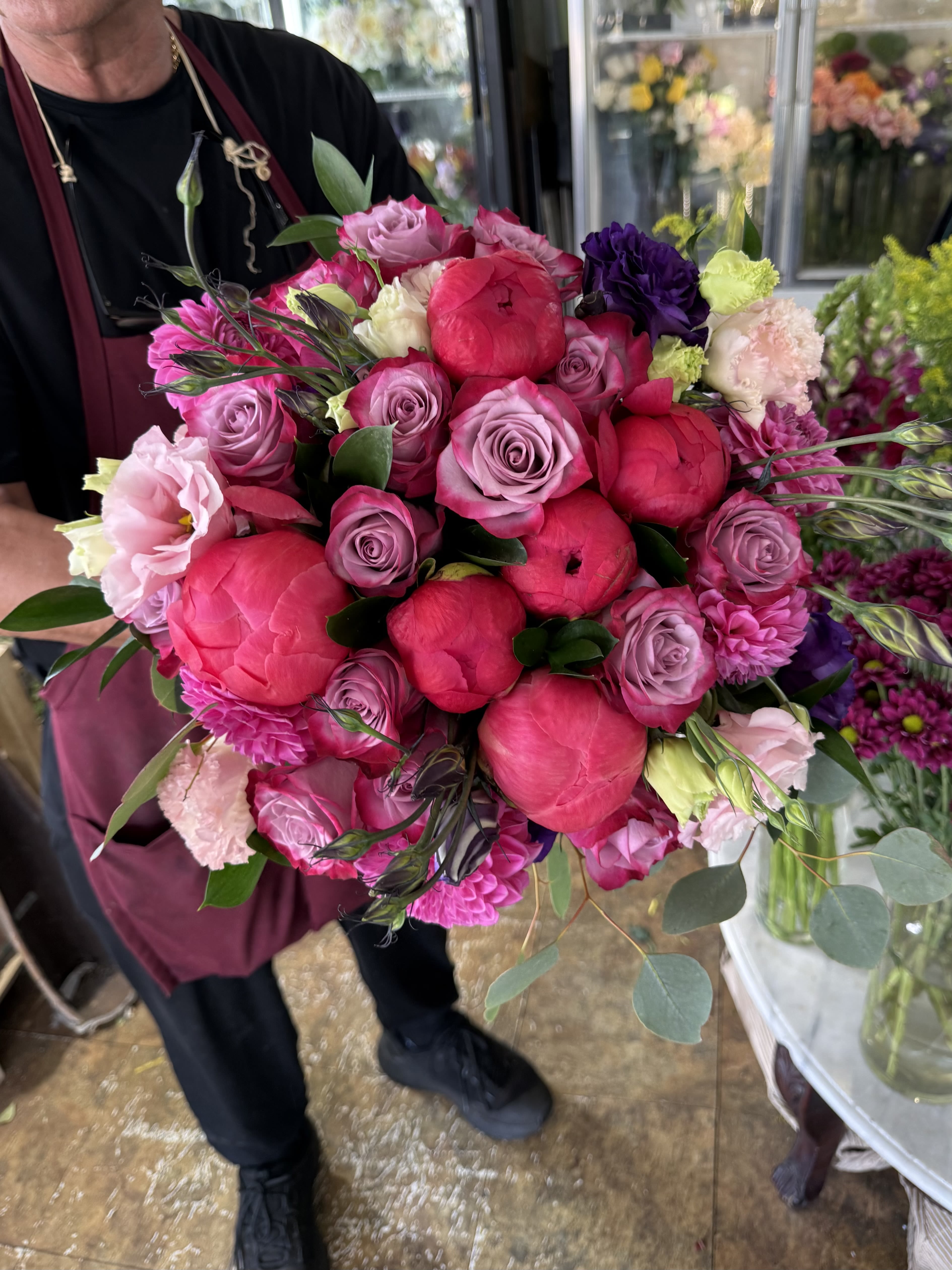 Peonies &amp; Provence - A lavish hand-tied bouquet where vibrant coral peonies meet elegant lavender roses, delicate lisianthus, and rich violent accents. Wrapped in crisp white paper, this bouquet is full of lush texture, romantic movement, and luxurious color-bringing together bold pinks, soft mauves, and deep plum tones in a design that feels both timeless and modern. Every stem is carefully chosen to create a bouquet that is vibrant, sophisticated, and unforgettable. 