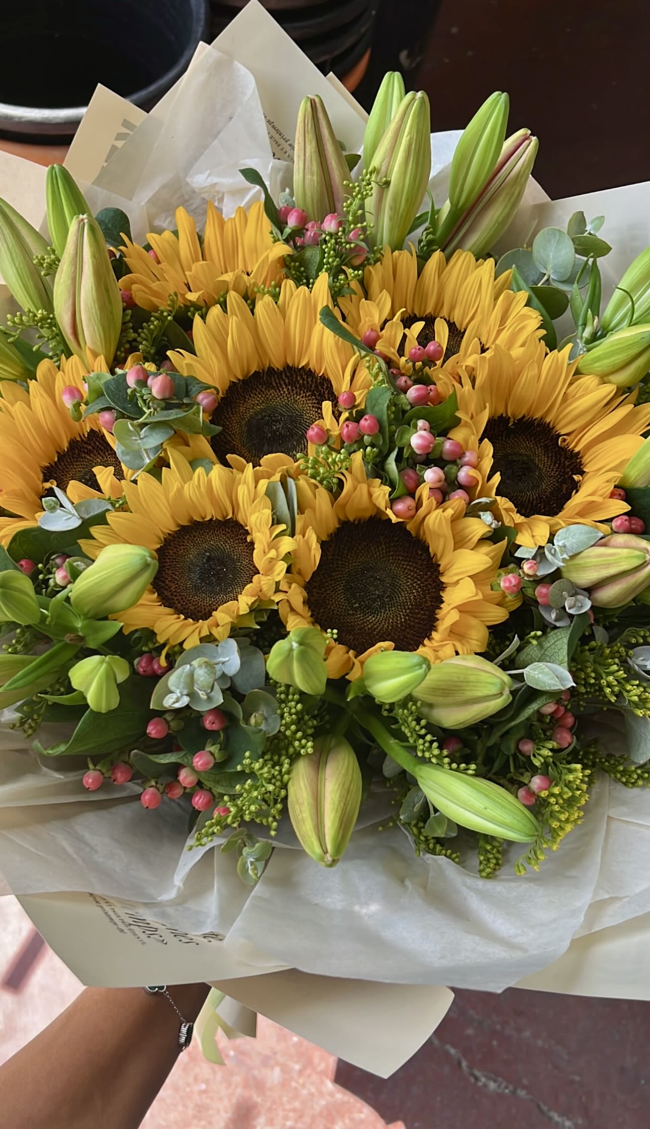 Hand-tied bouquet of yellow sunflowers with green lily buds and pink berries wrapped in cream paper.