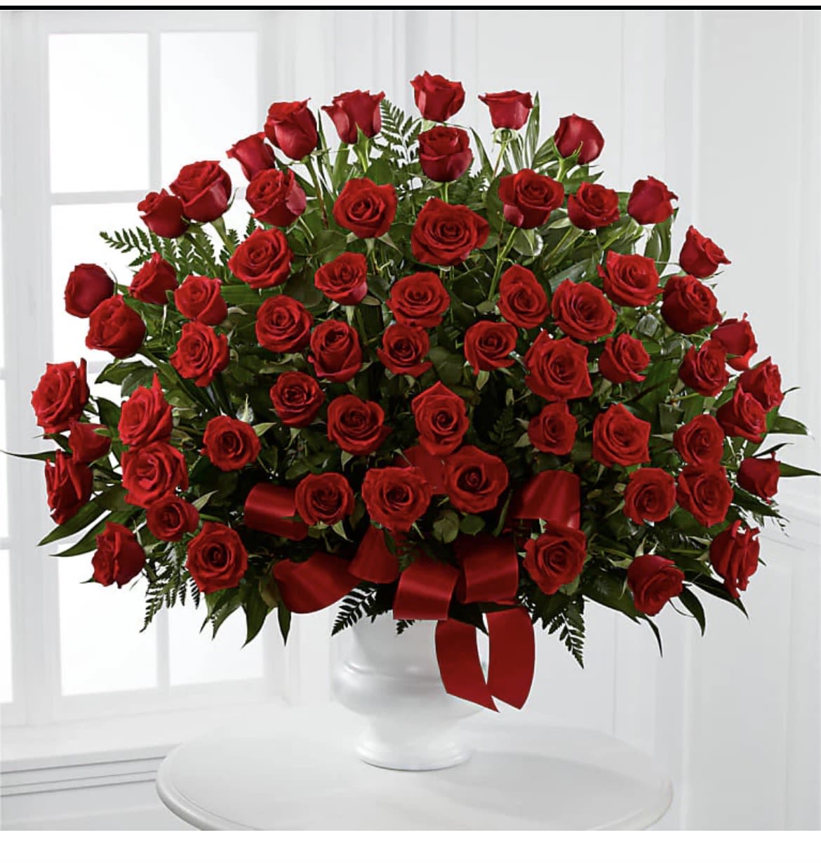 Red Romance  - A lush dome of deep red roses crowns a white ceramic vase, the kind of birthday, anniversary, or Valentine's bouquet that makes the person you're thinking of feel genuinely cherished. The rounded, densely packed arrangement reads formal and romantic-perfect for marking a milestone of love or for sending a respectful, elegant sympathy tribute when words fall short.  Each bloom is nestled against rich dark green foliage so the crimson petals read extra vivid; the presentation is finished with a wide red satin ribbon tied around the vase for a classic, luxurious touch. The white ceramic vessel keeps the focus on the flowers and cleanly contrasts the red and green, while the compact, abundant shape fills a room with visual warmth.  Ideal for anniversaries, Valentine's Day, romantic gestures, or solemn ceremonial moments, this arrangement is designed to convey deep feeling without fuss. Sized to make an impact on a table or mantle, it arrives ready to display; simply place in a cool spot away from direct sun and refresh the water every two days to keep the roses looking their best.