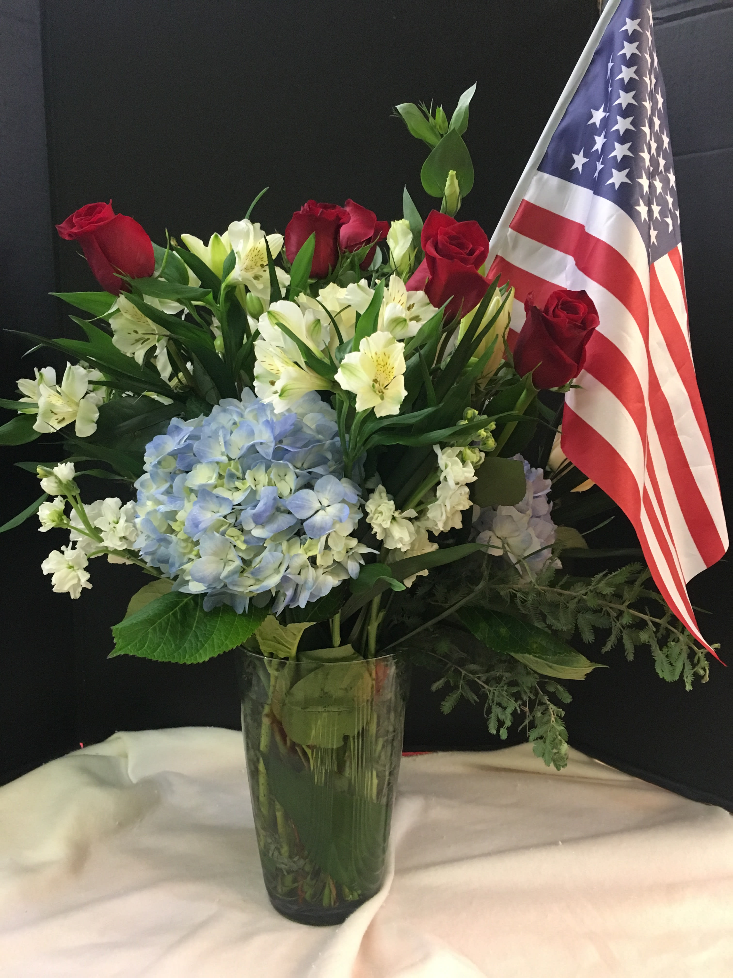 Red roses and white lilies with blue hydrangea in a glass vase beside an American flag