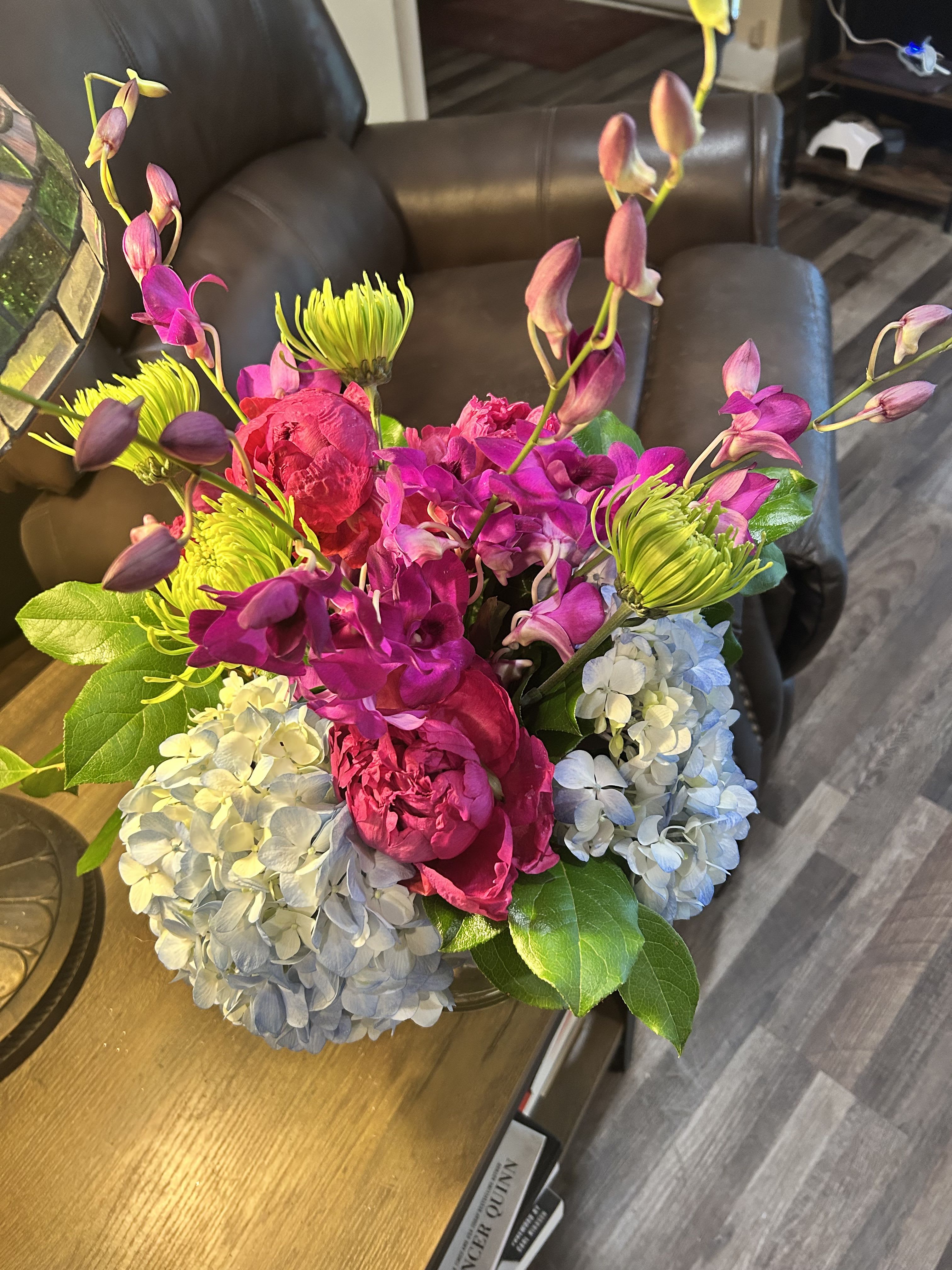 Colorful Cube 2 - Vivid magenta orchids crown a lush base of pale blue and white hydrangeas in this colorful cube flower arrangement, a joyful choice for a birthday bouquet, thank-you gift, or bright spring celebration. The contrast between saturated orchid blooms and soft hydrangea clusters makes the recipient feel celebrated and noticed the moment they see it.  Bright chartreuse spider mums weave energetic pops of color through glossy green foliage, while arching orchid stems add elegant movement above the dense, rounded composition. Delivered in a clear glass vase, the arrangement reads modern and upscale yet feels fresh and approachable-perfect for the person you want to surprise with something cheerful and stylish.  Designed to stand out on a table or desk, this arrangement suits everyday gifting or special moments when you want to say "you matter." Vase included for easy display; replace water and trim stems every few days to keep blooms looking their best.