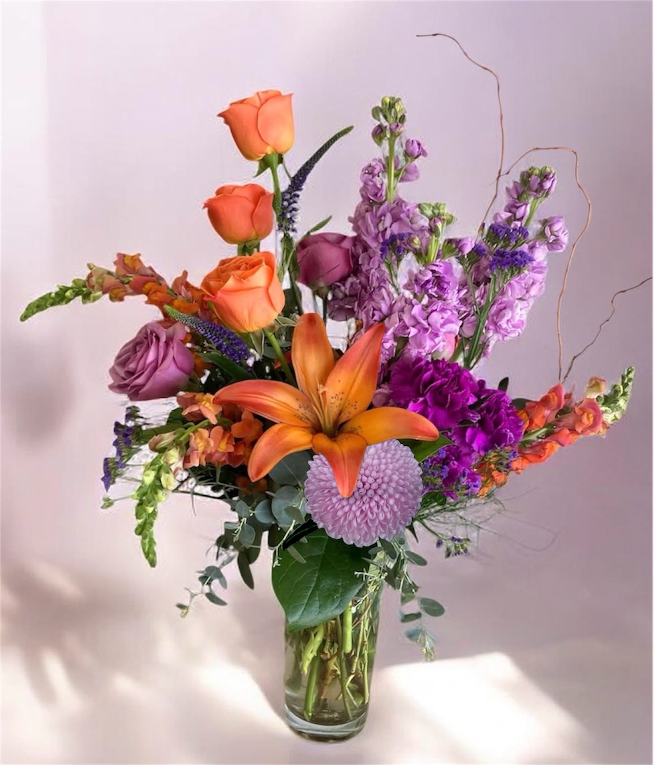 For The Queen - Bright orange lilies and snapdragons with purple roses, stock and carnations in a tall clear vase with curly branches, This purple and orange arrangement is a vibrant celebration for birthdays, congrats, cheerful surprises or for Mom, the queen herself.