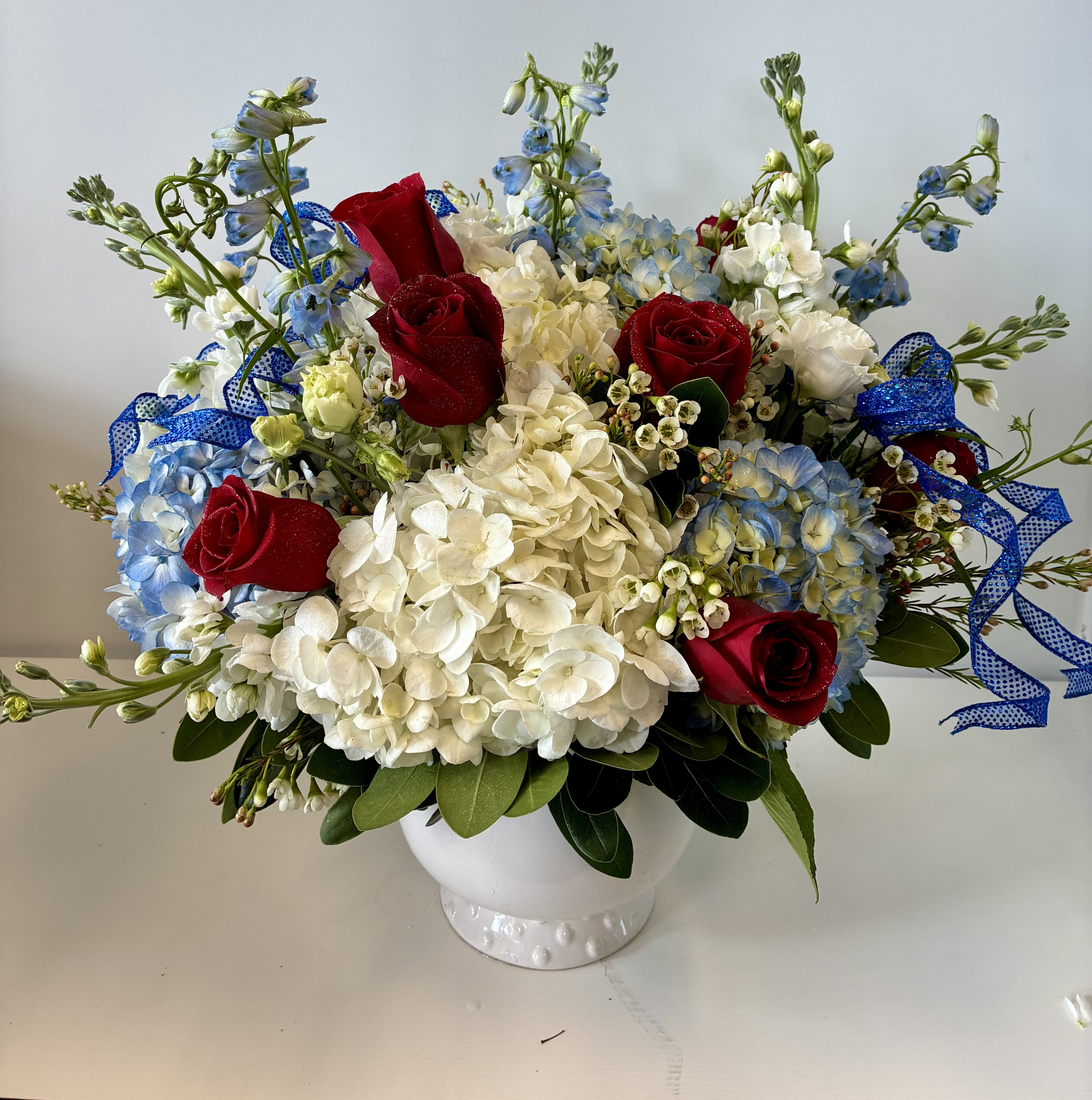 Sparkle &amp; Stems - Large white hydrangea clusters set a soft, cloud-like backdrop for deep red roses and airy blue stock in this rounded flower arrangement - a patriotic yet elegant choice for a Fourth of July table centerpiece.