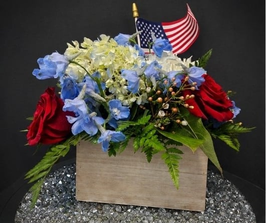 Happy 4th   - A bold display of patriotic pride, this Fourth of July flower arrangement is designed to make the recipient feel honored and celebrated. The centerpiece is a rectangular wooden box filled with a mix of red roses, pale blue blooms, and a generous cluster of white and cream hydrangea-like flowers-perfect as a Fourth of July centerpiece, Memorial Day tribute, or for any patriotic celebration where you want to say "thank you" or "we remember. "One small American flag add a ceremonial flourish that reads instantly festive and respectful.  Made to honor someone special, this arrangement suits veterans, hosts of a barbecue, or families gathering for a holiday observance-she'll feel seen and celebrated when these colors arrive. The wooden box keeps stems secure for simple placement; for best longevity, keep flowers in a cool spot away from direct sun and refresh water as needed.