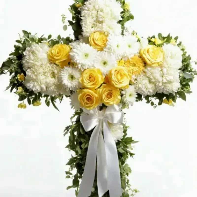 Sunshine Cross - A dignified sympathy cross designed to comfort and honor-a floral arrangement shaped as a large cross, perfect for memorial services or to send as a thoughtful sympathy tribute. The white, yellow and green palette brings together the reverence of white blooms with the warm, uplifting touch of yellow roses so the person you're remembering feels celebrated and gently remembered.  This arrangement pairs yellow roses with clusters of white chrysanthemums, daisies and other white blooms to form a dense, symmetrical cross with trailing greenery that softens the edges. A white ribbon is tied near the lower center for a formal finish; the composition is full and balanced, with strong floral coverage across the arms and center.  Choose this cross when you want a respectful, comforting tribute that reads both ceremonial and personal-the yellow roses add a hopeful note while the white flowers convey peace. Designed for easy placement at a service or graveside; professional delivery and care handling ensure the arrangement arrives fresh and appropriate for the occasion.