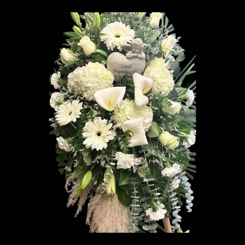 Angel spray - A serene, all-white memorial spray designed to honor and comfort - perfect as a sympathy floral tribute or for a funeral service. This standing arrangement communicates sympathy and reverence for the person you're remembering, with a formal upright shape that reads respectful from a distance and close up.  Dense clusters of white Asiatic lilies, gerbera daisies, hydrangeas, calla lilies and carnations are layered with abundant green foliage for depth and texture; smaller filler blooms soften edges and create a full, polished silhouette. A small memorial heart plaque and a delicate angel figurine are placed near the center, and the spray is displayed on a standing easel for easy placement at a service or graveside.  Give this as a meaningful expression of sympathy to help the bereaved feel seen and supported. The arrangement's size and upright design make it suitable for chapel display or as a graveside tribute; please mention any personalization needs when ordering so we can position the memorial plaque or figurine as requested.
