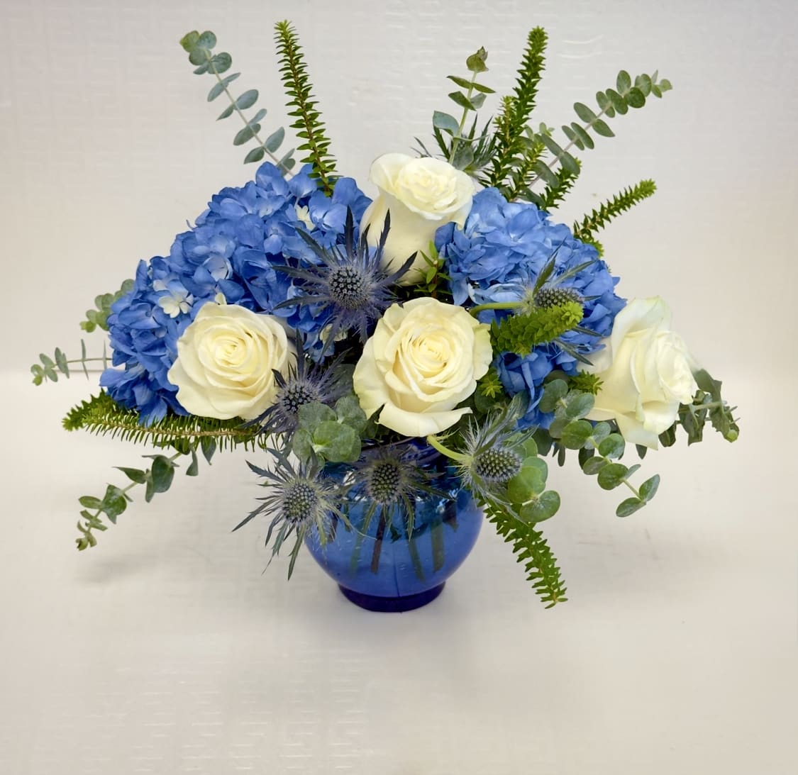 Blue Serenity Bouquet - A sophisticated blend of vibrant blue hydrangeas, creamy white roses, and striking blue thistle accented with fresh eucalyptus and lush greenery. Designed in a rich blue vase. A timeless design that conveys peace, and heartfelt sentiment, making it perfect for birthdays, sympathy, get well wishes, thank you gifts or simply brightening someone's day.
