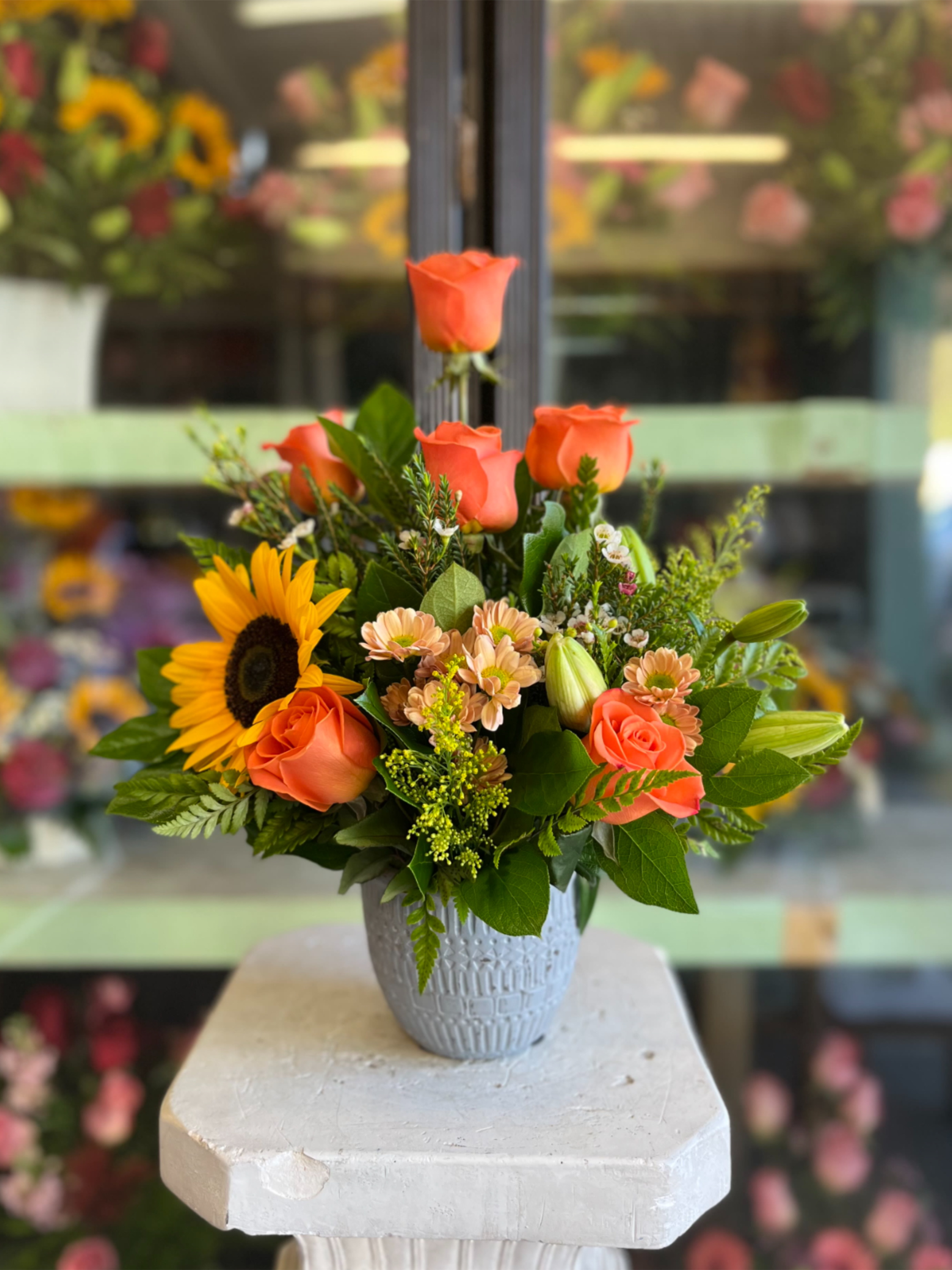 Corral Rose Sunflower Small Arrangement - Bright coral roses and a single sunny sunflower mingle with soft peach spray chrysanthemums, pale lily buds, and mixed greenery in a textured gray ceramic vase-an upbeat flower arrangement perfect for birthdays, thank-you gifts, or a cheerful just-because delivery. The compact, garden-style design feels fresh and celebratory, sending a warm, sunlit message to the person you're thinking of.