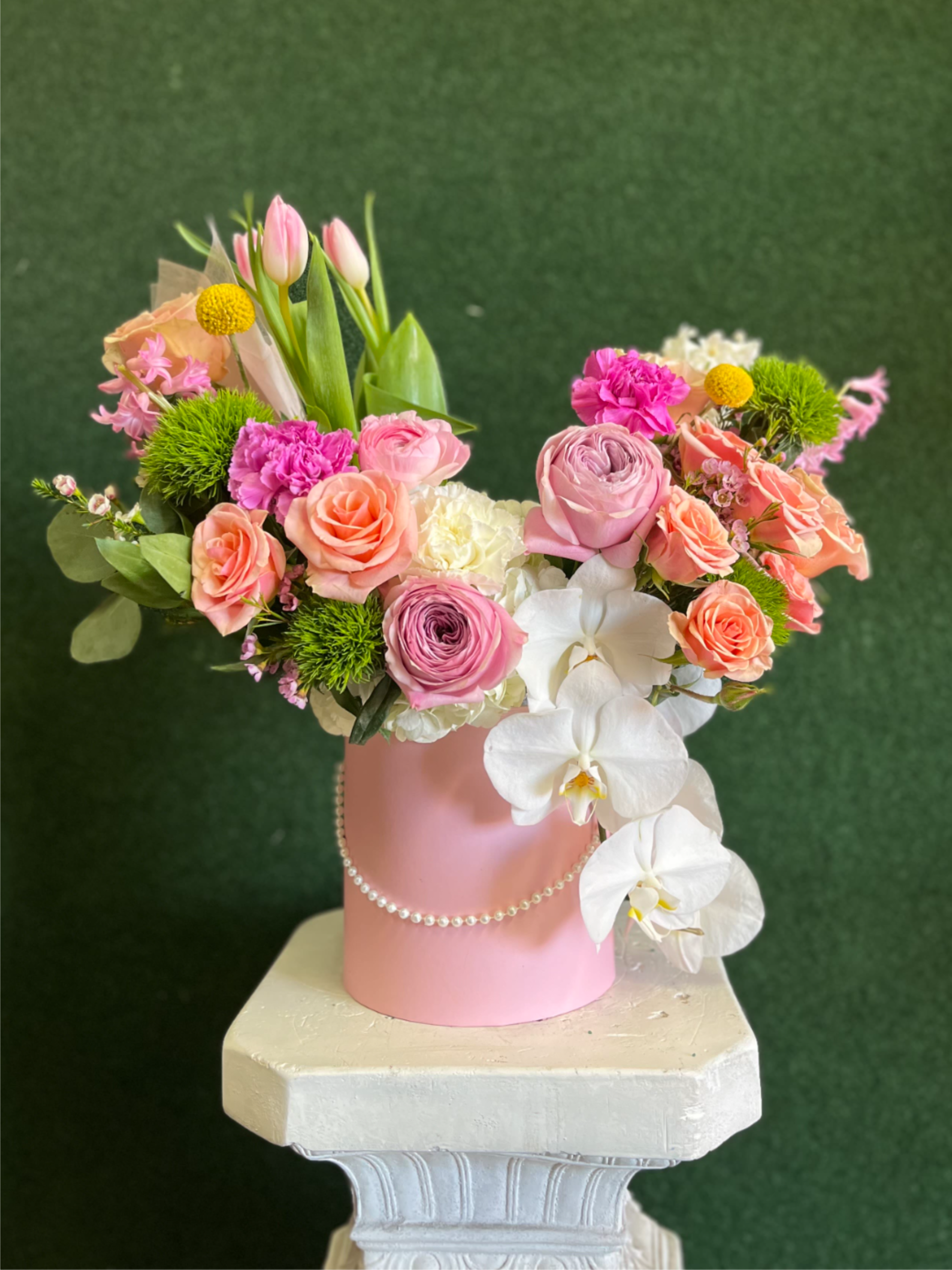 Pink Tulip And Rose Box Arrangement - A lush, rounded flower arrangement of pink and peach roses, white orchids, pink tulips, hydrangeas and carnations designed for birthdays, anniversaries or wedding celebrations - the feminine palette and bright green textural accents create a polished, romantic impression the recipient will feel truly celebrated by. Arranged in a matte pink cylindrical vase trimmed with a pearl garland and presented on a pedestal-style display, this composition offers abundant texture and cascading orchids for visual movement and lasting presence.
