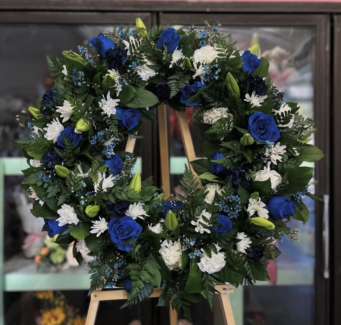 Blue Sympathy Floral Arrangement - The wreath is built on a sturdy wreath frame with dense green foliage, punctuated by darker blue blooms, small blue filler flowers, and several lily buds for texture and depth. Arranged to be viewed from the front, the piece comes with a wooden easel and a custom ribbon, presented at a scale suitable for viewing at a service or graveside.  A meaningful way to express condolences, this wreath will help the recipient feel remembered and respected during a difficult time. For delivery and sizing details or to request a specific ribbon message, please contact us when placing your order.