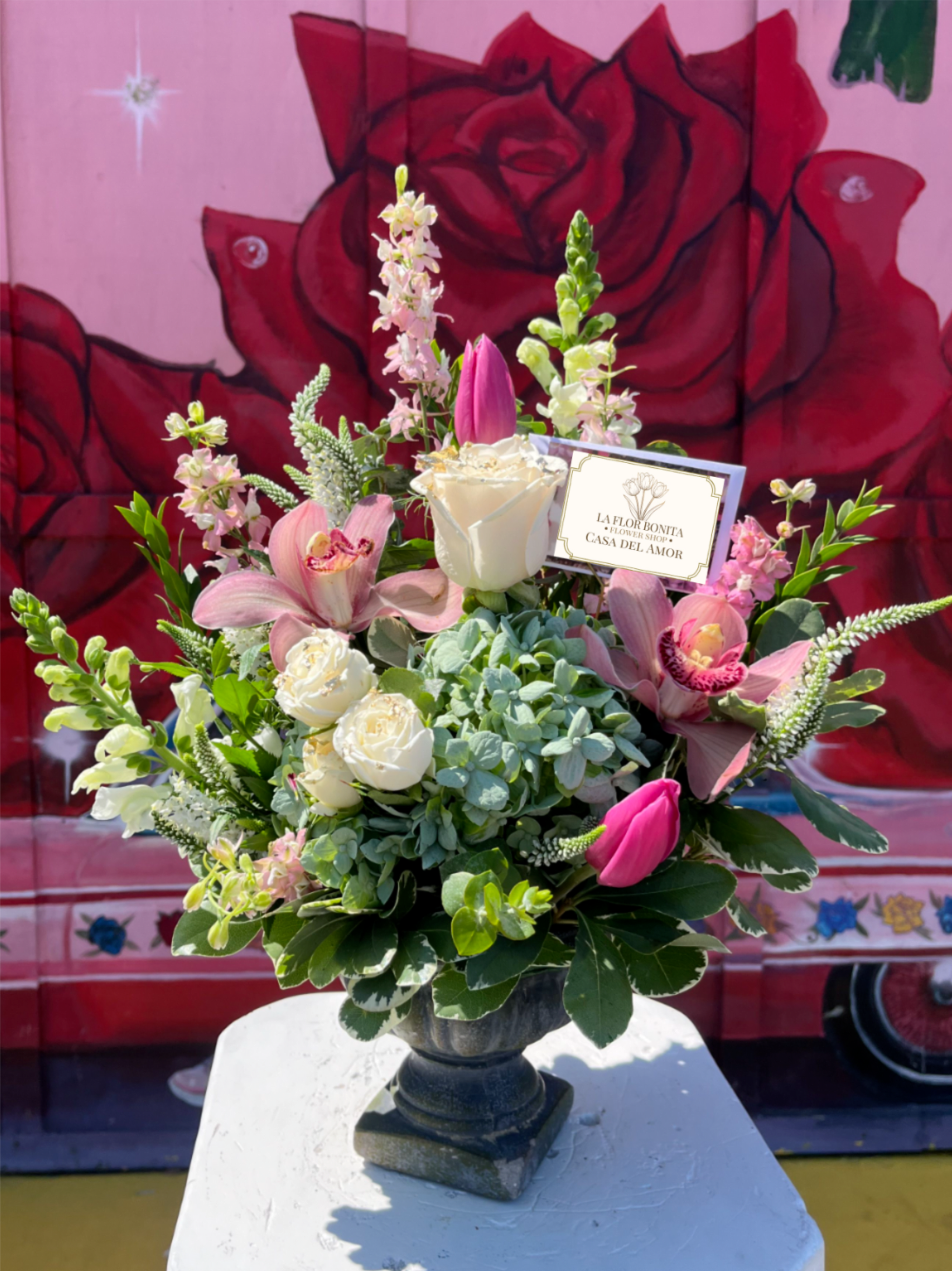 Snapdragon And Pink Orchid Arrangement - Soft blush orchids and upright snapdragons rise above a lush base of white roses, pink tulips, and blue‑green hydrangea in a dark pedestal vase-an elegant flower arrangement perfect for an anniversary bouquet or a romantic birthday gift. The layered, garden‑style composition and the tucked greeting card make the recipient feel celebrated and truly seen.  Rendered with the refined restraint of a Roman garden-timeless, graceful, and ceremonious-this arrangement pairs sculptural blooms with verdant foliage for a luxurious presence in any room. Presented in a ceramic urn-style vase with a florist card, it arrives ready to display and enjoy.